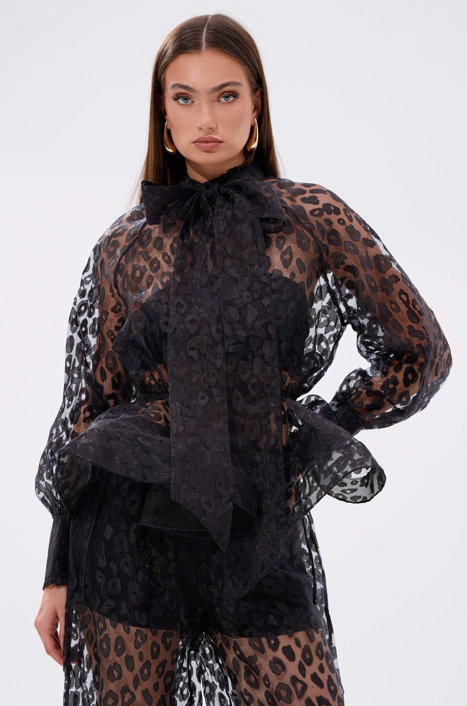 A woman models the CHEETAH SISTA TIE FRONT BLOUSE, a sheer black and brown cheetah print top featuring a large tie front bow at the neck. She pairs it with matching shorts and gold hoop earrings, standing against a plain light background.