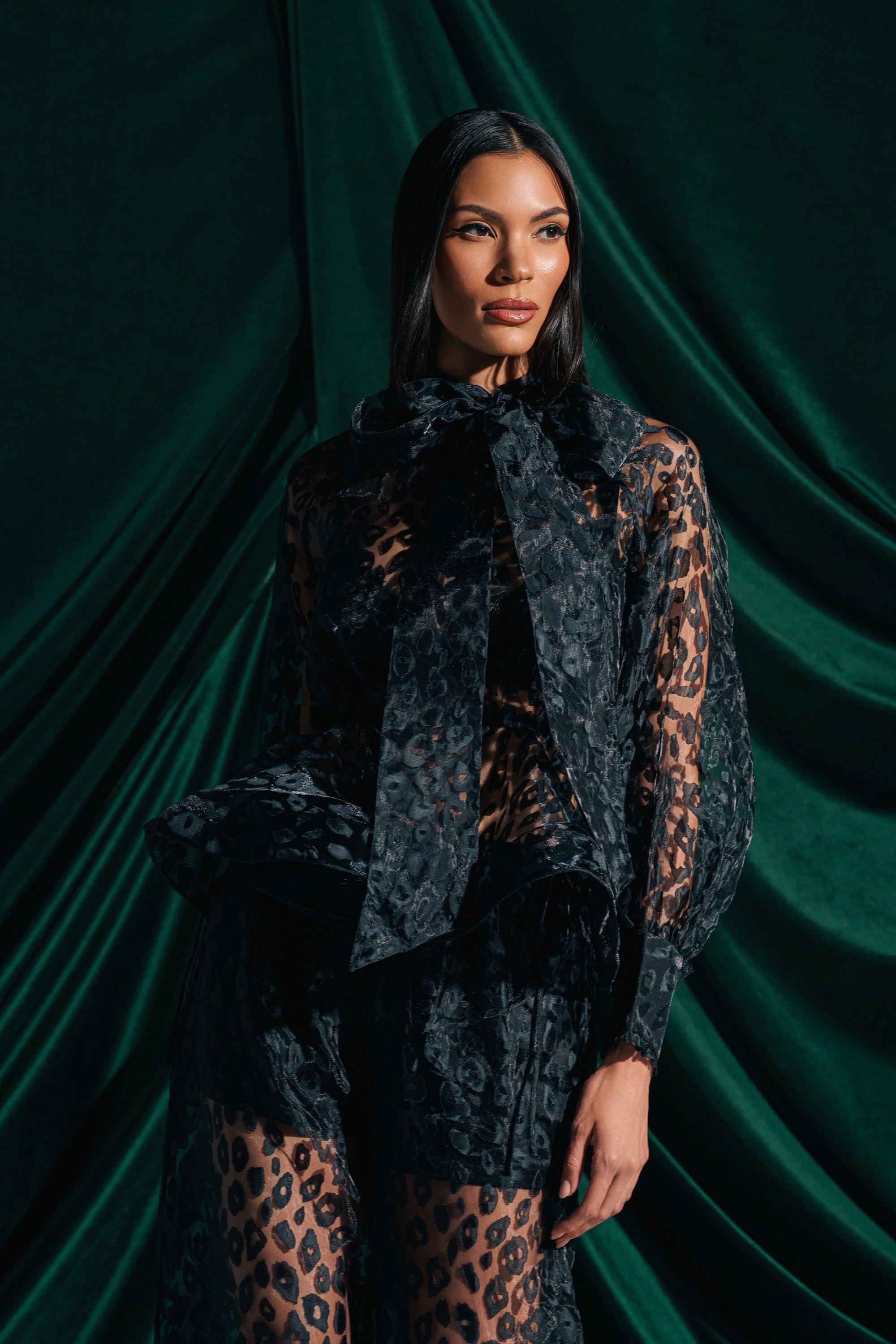A woman poses confidently before dark green fabric, wearing the CHEETAH SISTA TIE FRONT BLOUSE— a sheer black cheetah print top with an oversized neck bow—paired with matching pants. Her long hair is sleek and straight.