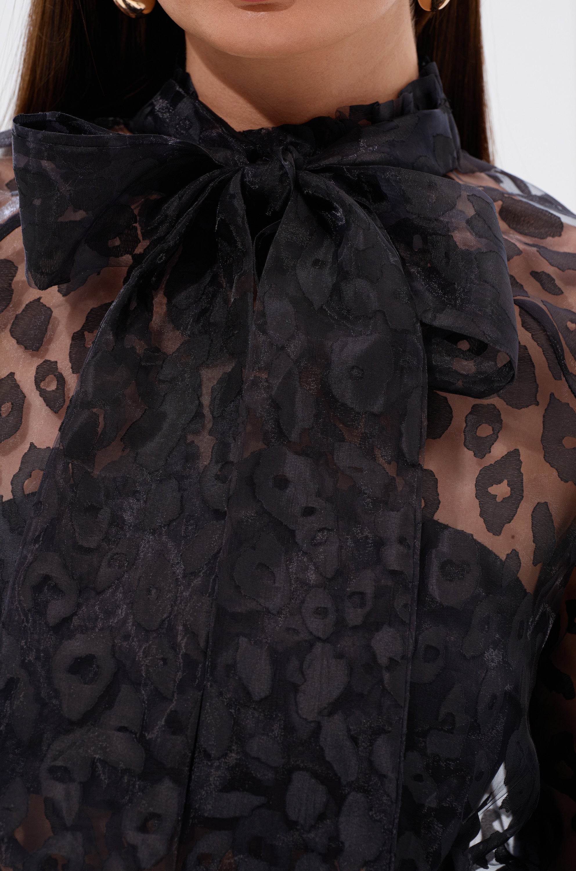 Close-up of a woman wearing the CHEETAH SISTA TIE FRONT BLOUSE—a sheer black blouse with a delicate floral pattern and a large bow at the neck. The see-through fabric adds allure, paired with gold hoop earrings for a chic finish.
