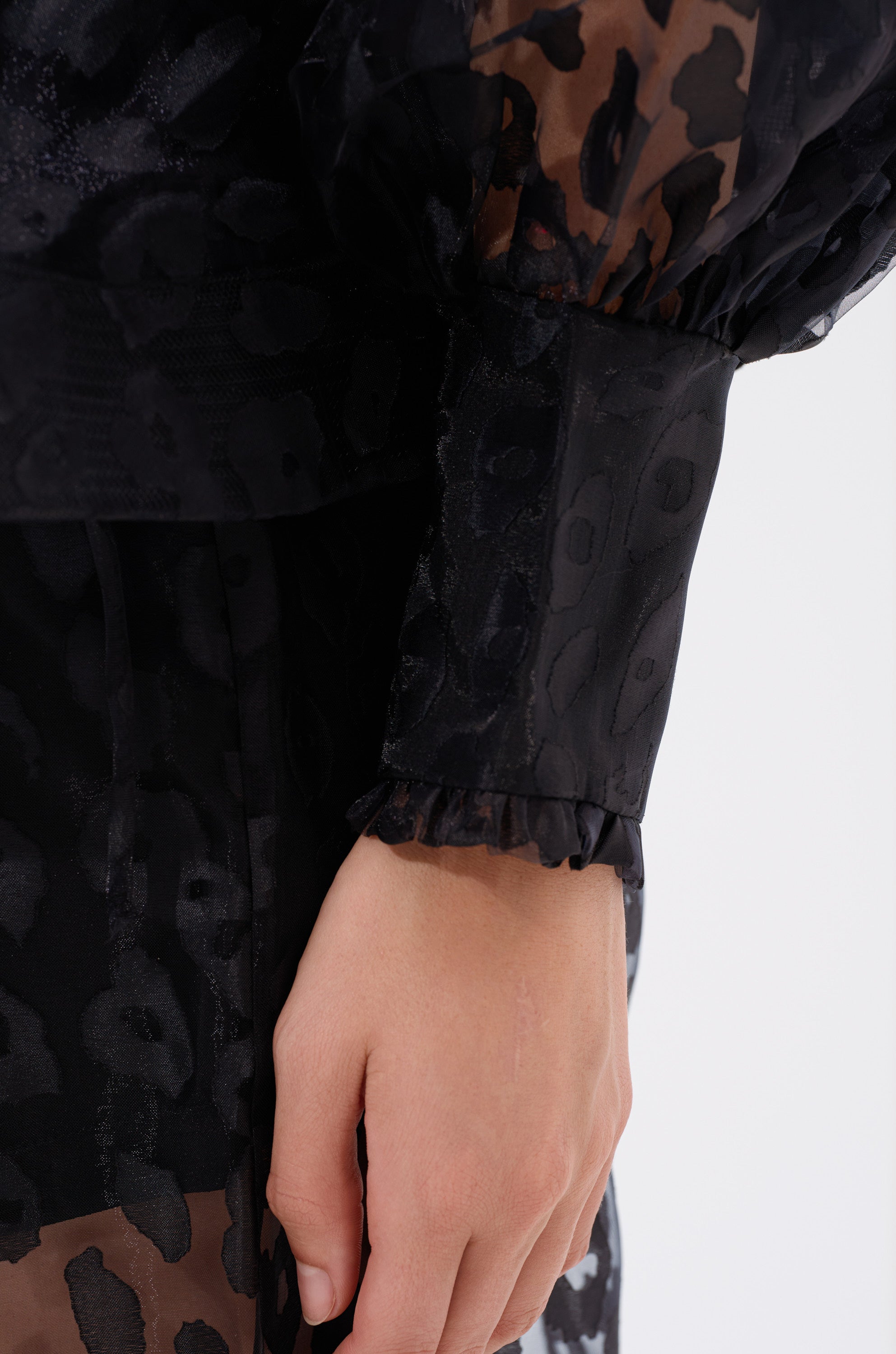 A close-up of a hand by the side, showing the sheer, ruffled cuff of the CHEETAH SISTA TIE FRONT BLOUSE. Its translucent black cheetah print fabric adds an elegant touch to the statement sleeve.