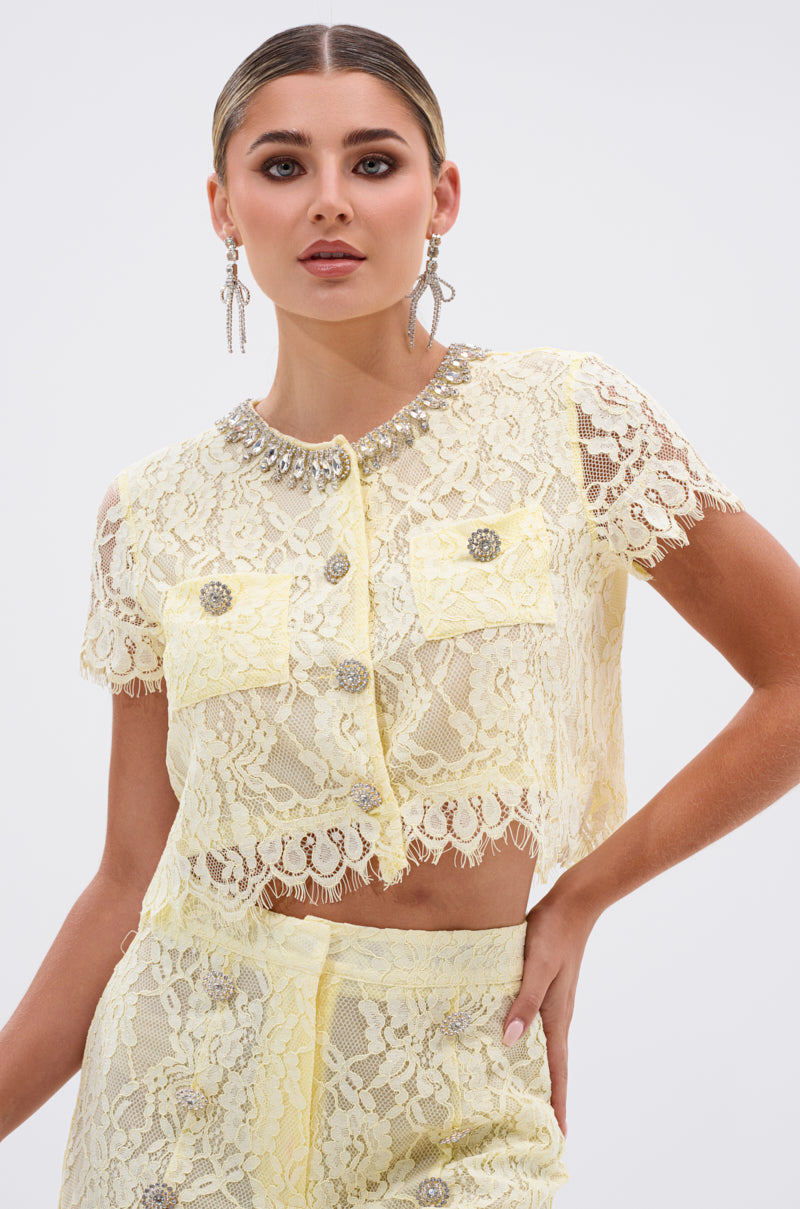 A woman wears the LEAH LACE BUTTON DOWN BLOUSE IN BABY YELLOW with matching high-waisted lace shorts, featuring decorative buttons and a jeweled collar. She styles it with slicked-back hair and dangling earrings.