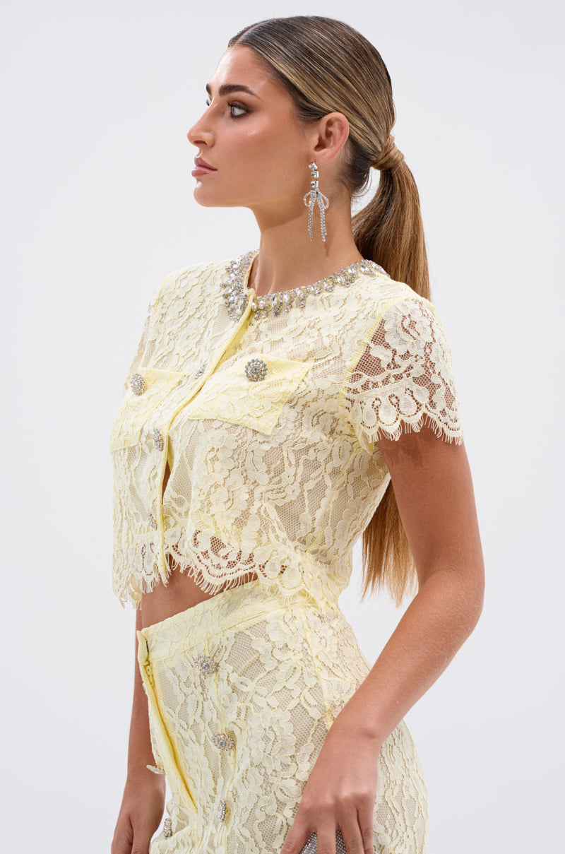 Woman with long, straight hair in a low ponytail wears the LEAH LACE BUTTON DOWN BLOUSE IN BABY YELLOW, featuring sheer floral lace and jeweled buttons, paired with a matching skirt. She is shown in profile against a light background.