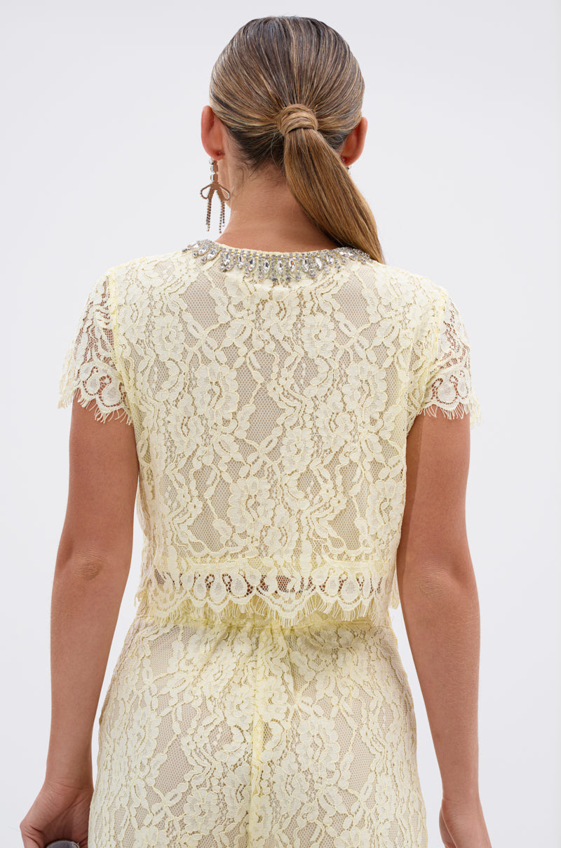 A person with straight light brown hair in a low ponytail is shown from the back, wearing the LEAH LACE BUTTON DOWN BLOUSE IN BABY YELLOW—a sheer floral lace top with short sleeves and a jeweled neckline.