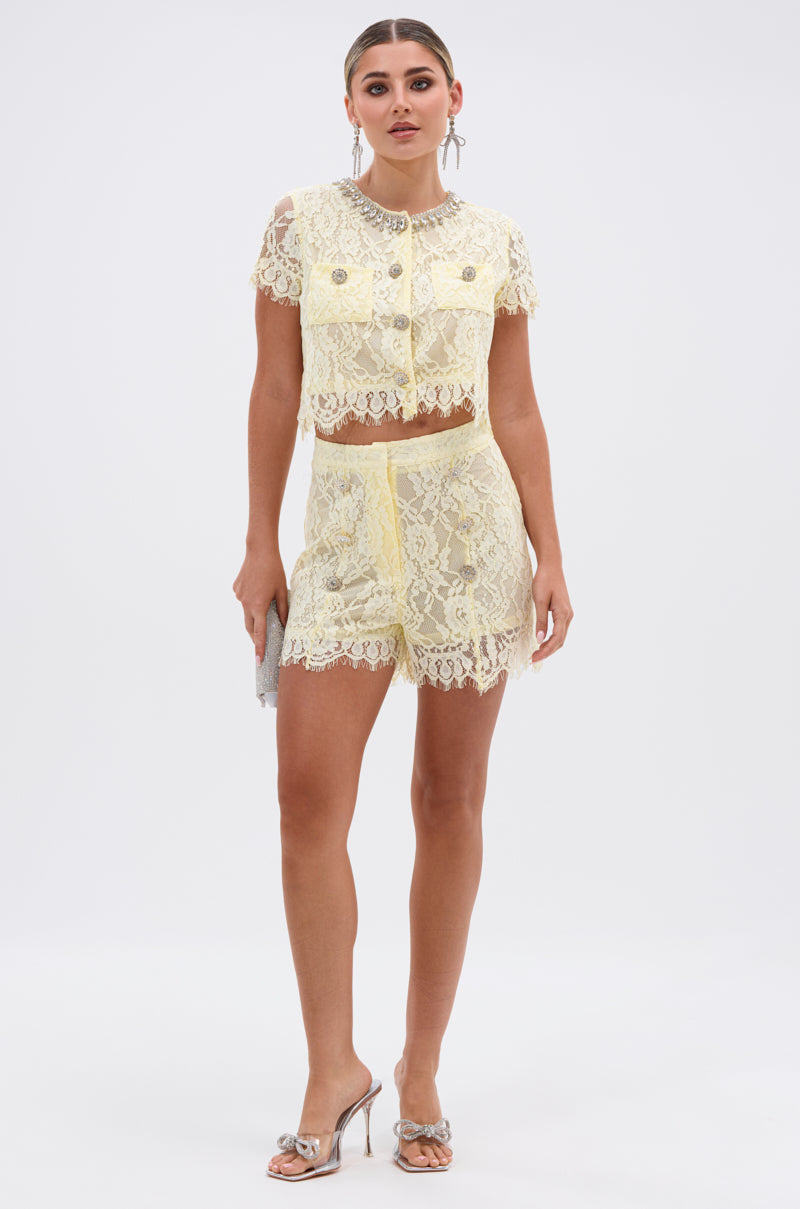 A woman poses against a white background in the LEAH LACE BUTTON DOWN BLOUSE IN BABY YELLOW paired with matching high-waisted shorts, accessorized with a silver clutch and silver heels for a chic look.