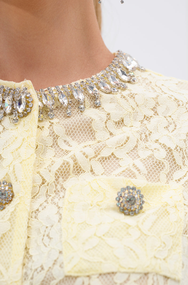 Close-up of a person wearing the LEAH LACE BUTTON DOWN BLOUSE IN BABY YELLOW, a sheer floral lace blouse featuring crystal embellishments at the neckline and sparkling beaded buttons on lace pockets.