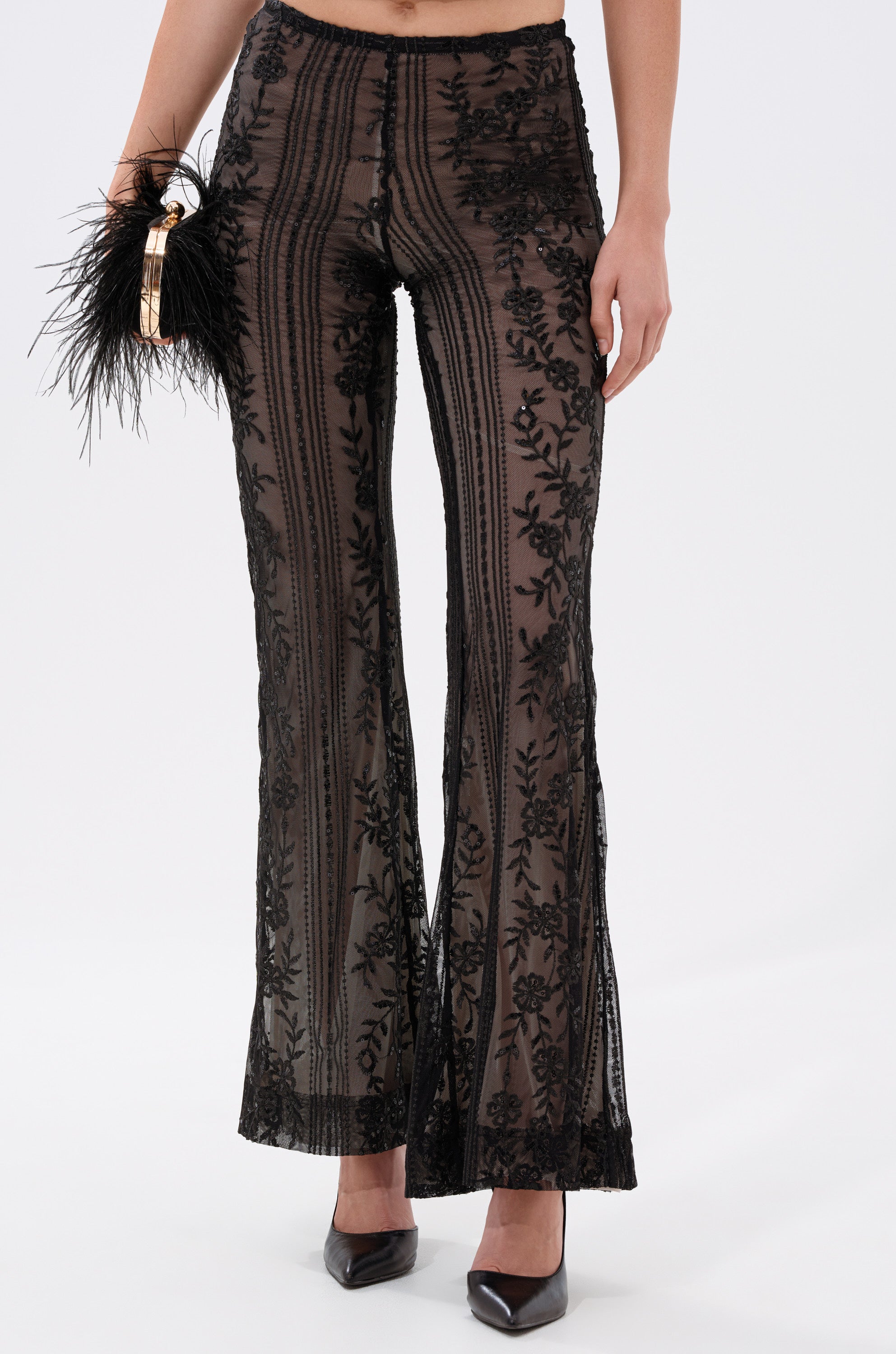 Wearing DEZI LACE TROUSER with sheer black mesh and floral patterns, paired with a black feathered clutch and black pointed-toe heels for an elegant, stylish look. 0
