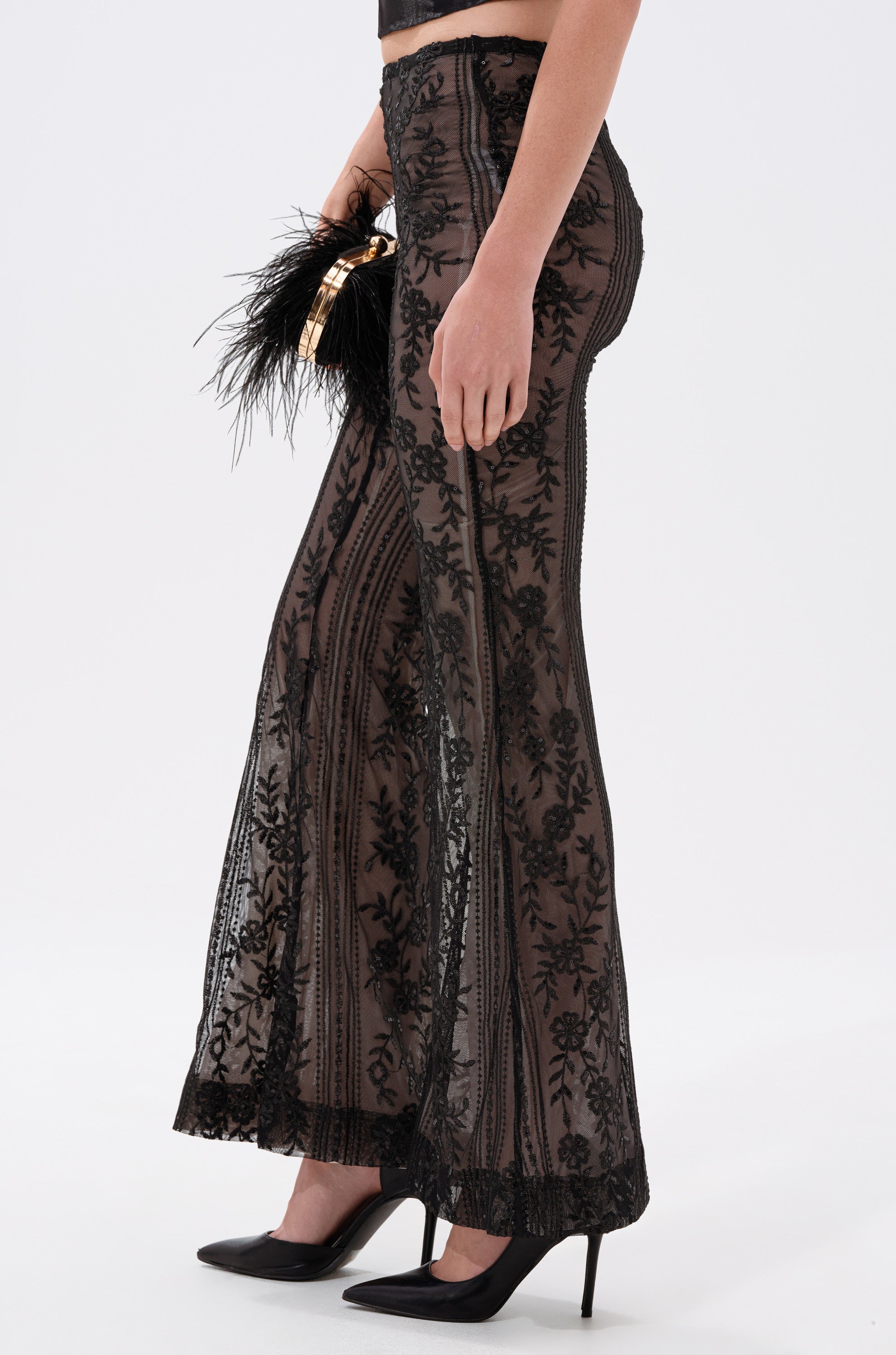 A person wearing the DEZI LACE TROUSER—black trousers with sequin-embellished floral lace and sheer mesh accents. 2