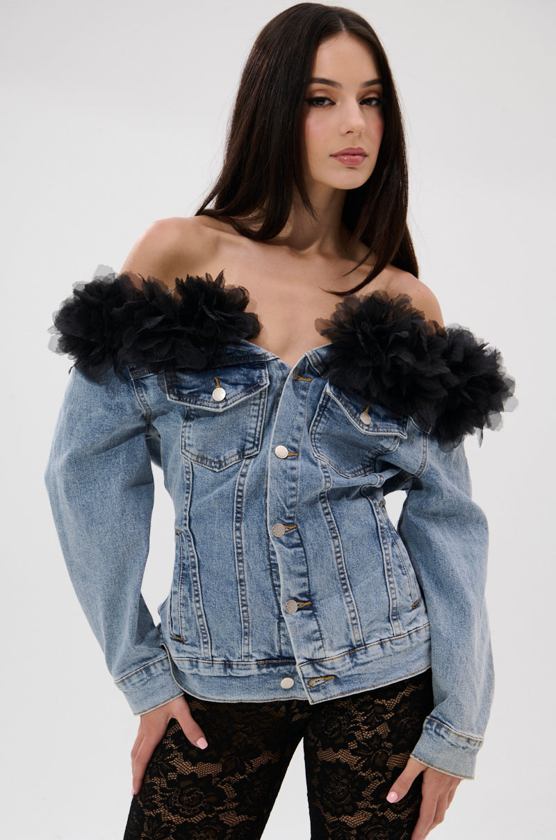 A woman with long dark hair models the DANA OFF THE SHOULDER RUFFLE DETAIL DENIM TOP, featuring large floral tulle ruffles, paired with black lace pants against a plain light background.