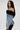 Woman with long dark hair wears the DANA OFF THE SHOULDER RUFFLE DETAIL DENIM TOP, featuring black tulle trim and a lace-up back, paired with black lace pants, standing sideways against a plain light background.