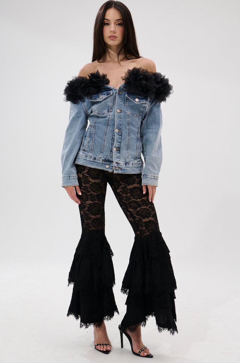 A woman poses against a plain background wearing the DANA OFF THE SHOULDER RUFFLE DETAIL DENIM TOP, featuring lace-up back and black tulle trim, paired with black lace ruffled flared pants and black high-heeled sandals.