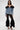 A woman poses against a plain background wearing the DANA OFF THE SHOULDER RUFFLE DETAIL DENIM TOP, featuring lace-up back and black tulle trim, paired with black lace ruffled flared pants and black high-heeled sandals.
