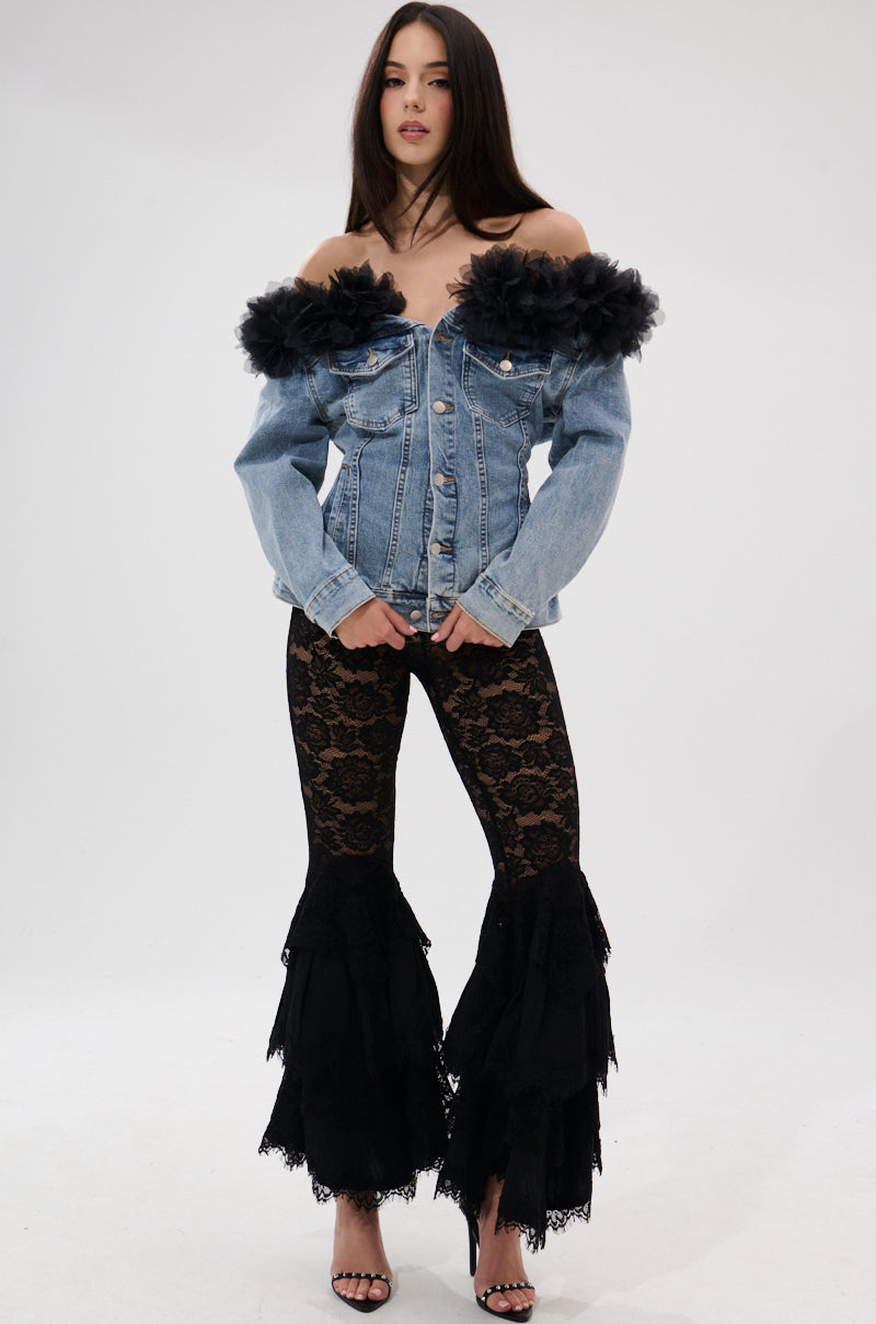 A woman poses against a white background in the DANA OFF THE SHOULDER RUFFLE DETAIL DENIM TOP, paired with black lace tiered flared pants and high-heeled sandals. The outfit features ruffled accents and a chic lace-up back.