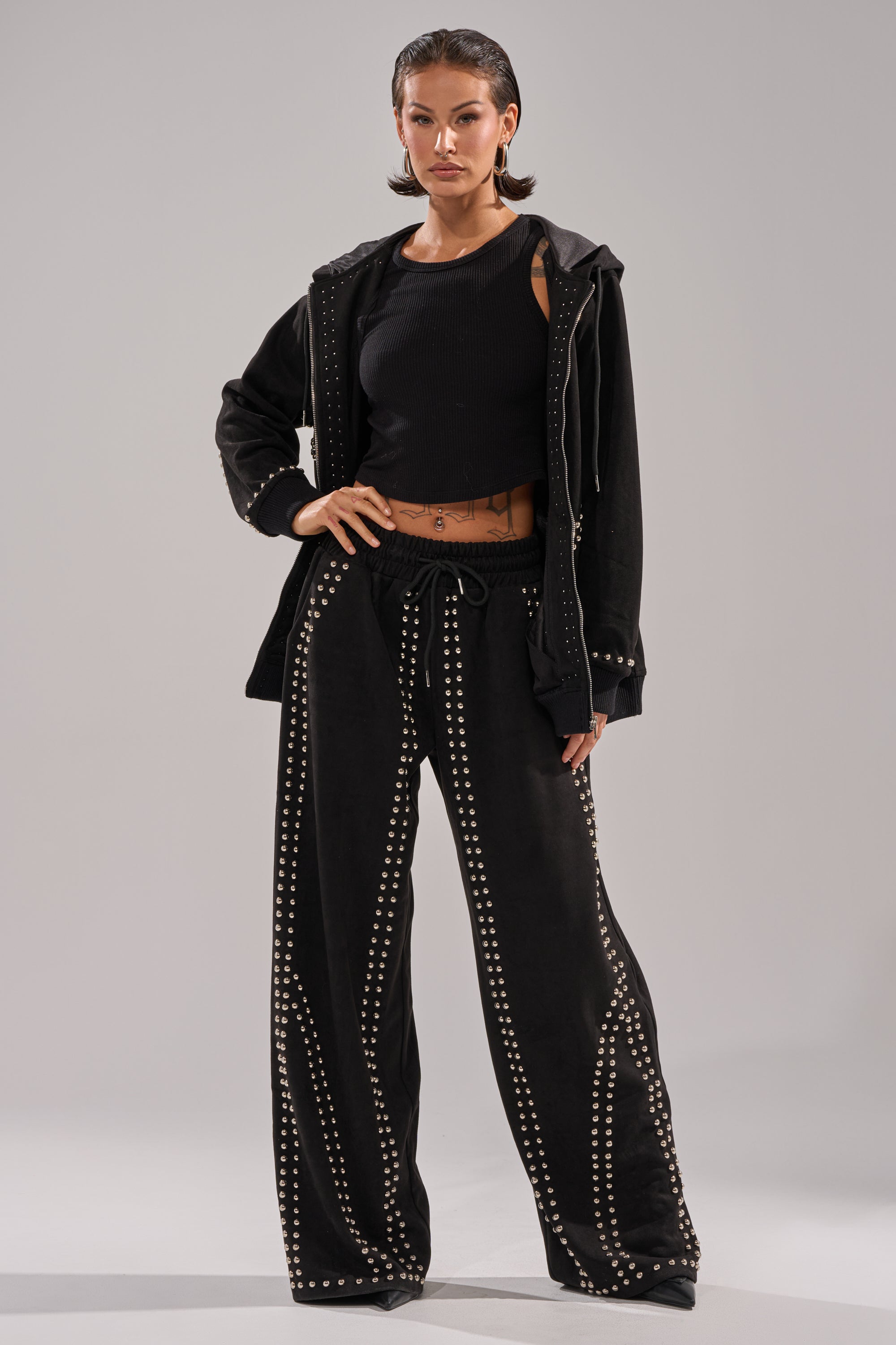 A woman poses confidently in the ASHLEY STUDDED WIDE LEG JOGGER, paired with a black sleeveless top and matching jacket, standing with one hand in her pocket against a plain light gray background.