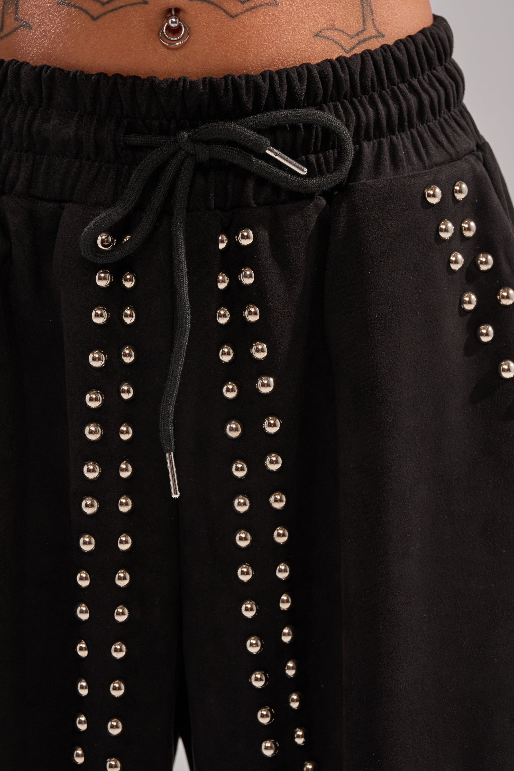 Close-up of a person wearing the ASHLEY STUDDED WIDE LEG JOGGER, featuring black fabric and vertical metallic accents. The high-rise fit reveals a tattoo and belly button piercing on the model’s lower torso.