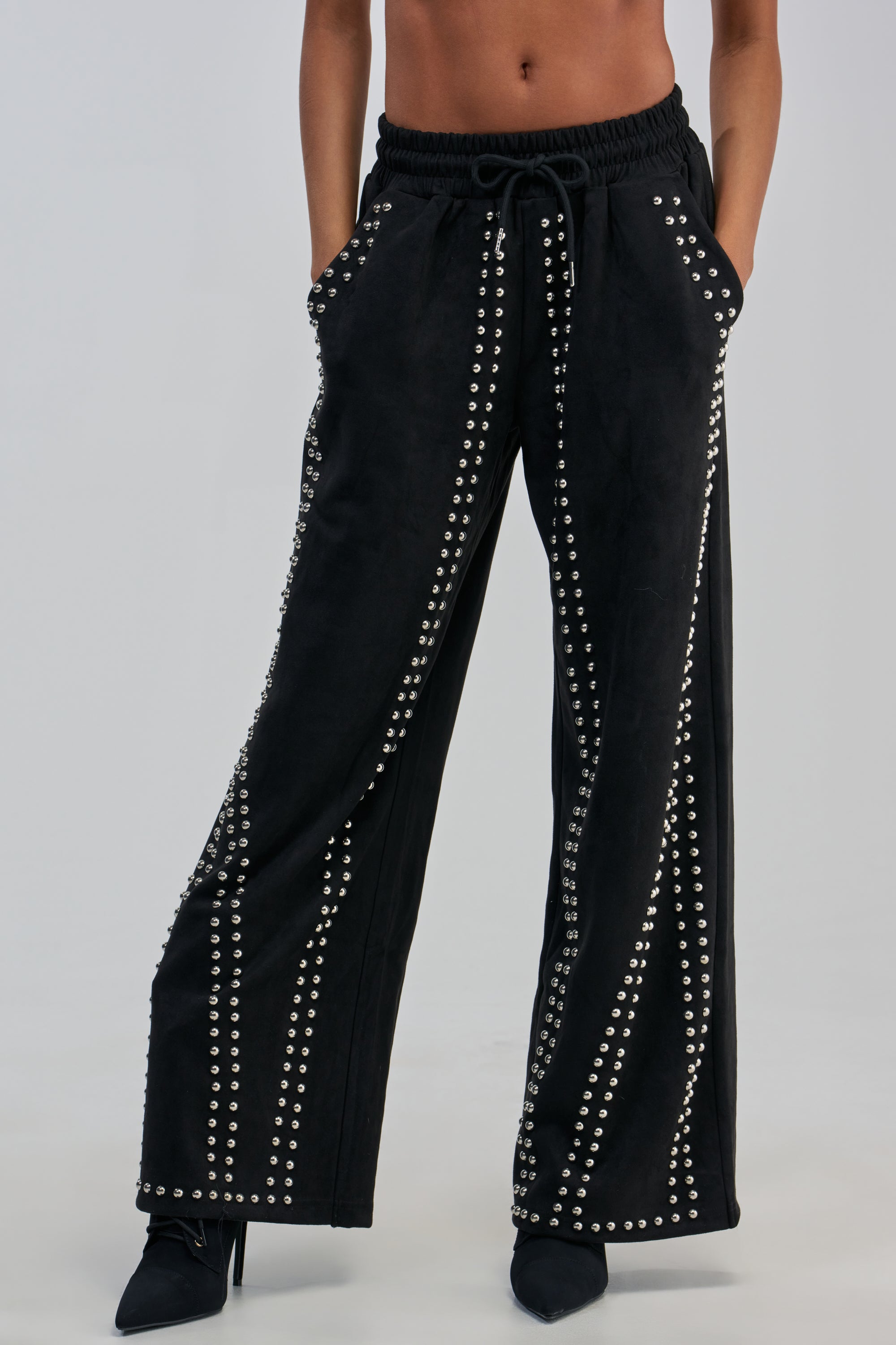 A person wearing the ASHLEY STUDDED WIDE LEG JOGGER, featuring silver studs in vertical lines, with hands in pockets and paired with black pointed-toe heels, shown from midriff to feet.