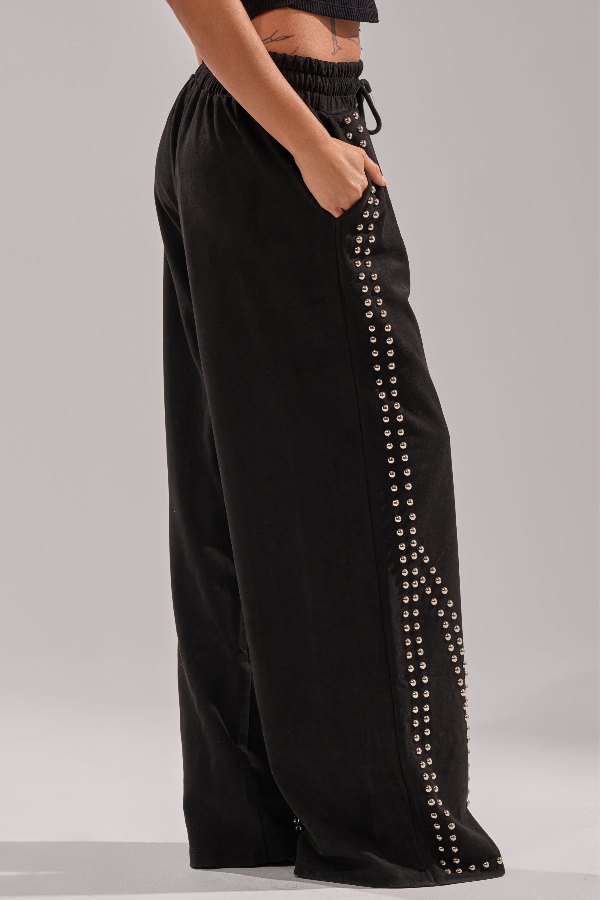 Wearing the ASHLEY STUDDED WIDE LEG JOGGER with silver studs along the sides and pockets, a person stands with one hand in the pocket, showing a small tattoo on their exposed midriff.