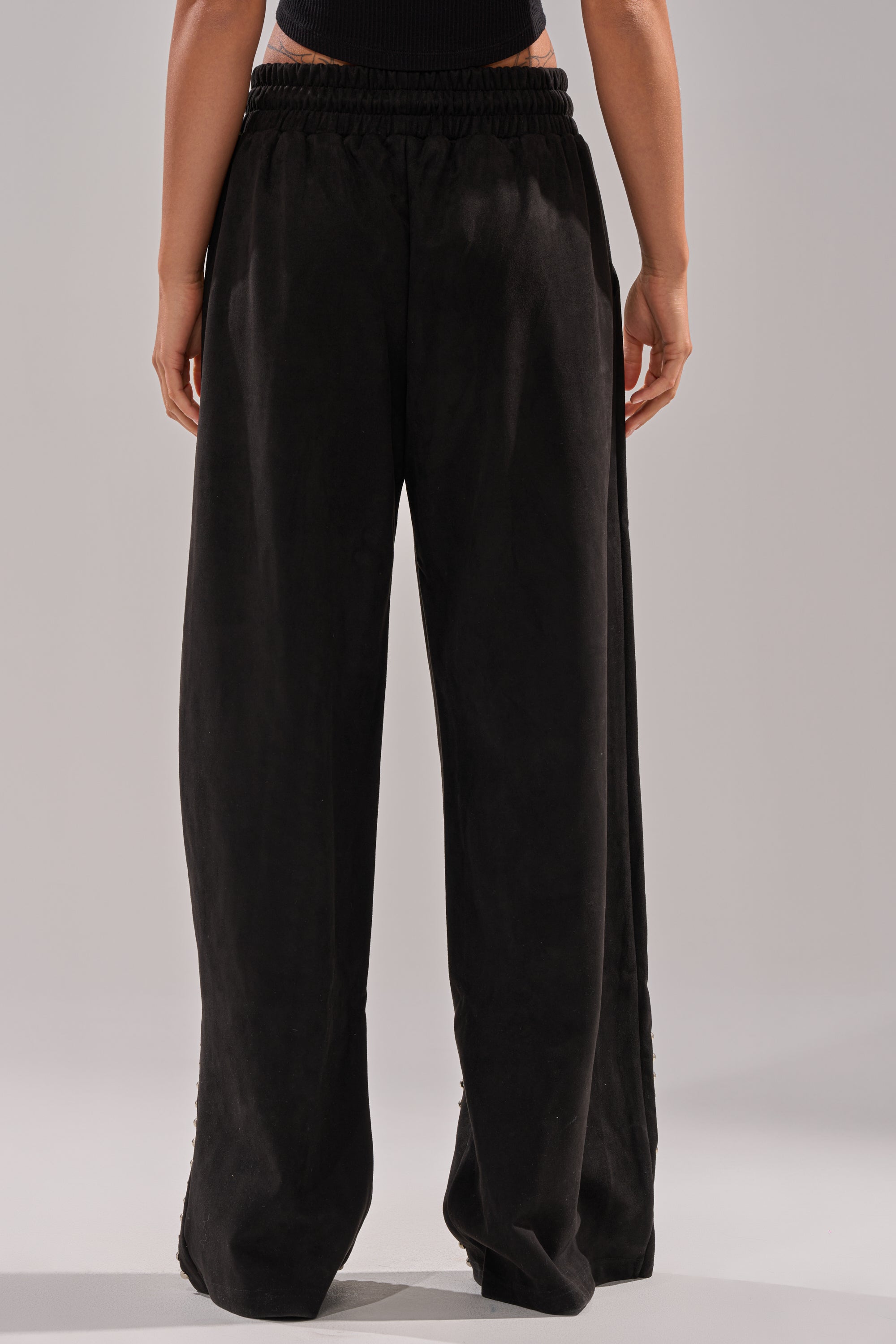 A person stands facing away, wearing the ASHLEY STUDDED WIDE LEG JOGGER in black with an elastic waistband and a black top against a plain, light background.