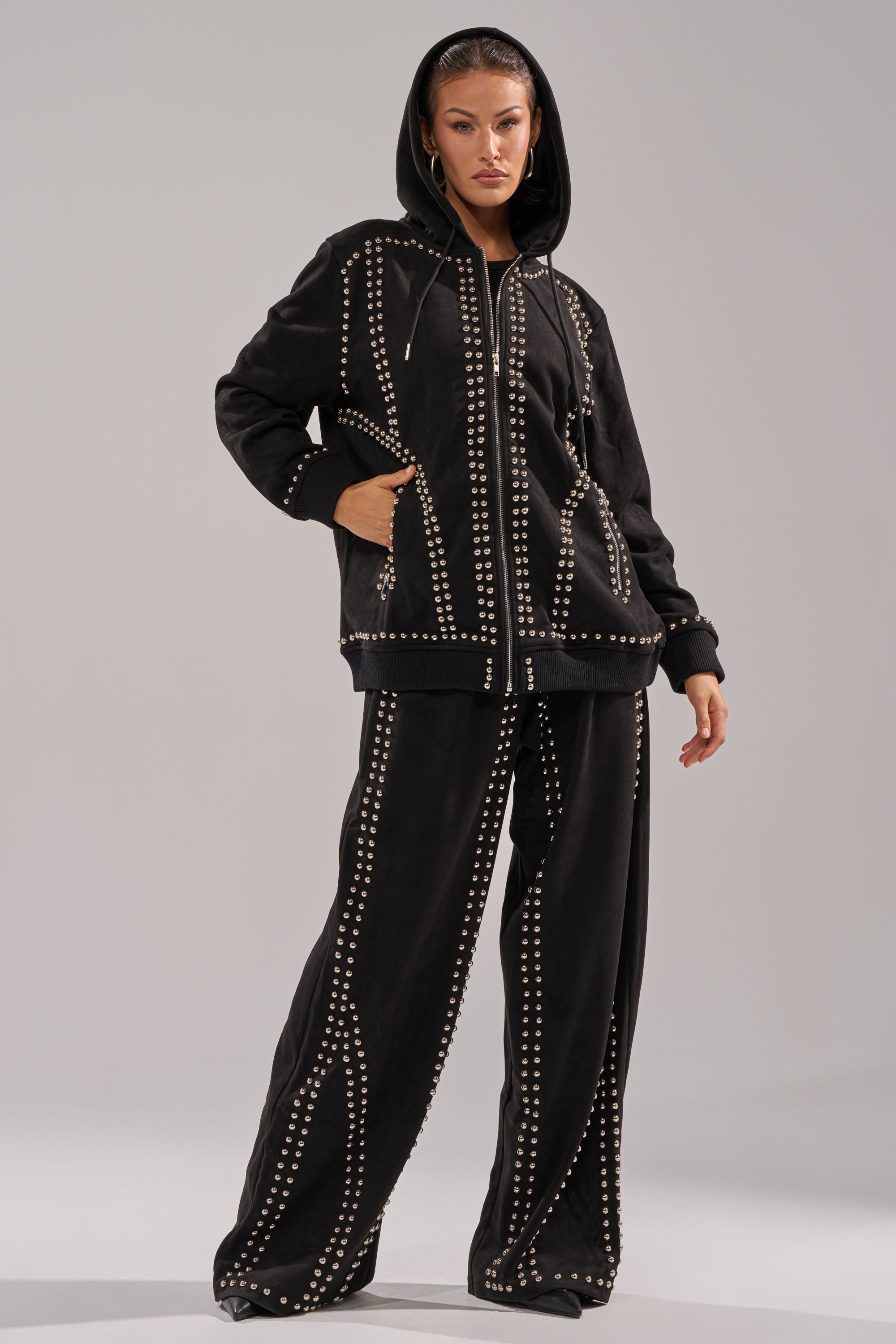 A woman wears the ASHLEY STUDDED WIDE LEG JOGGER and a matching black hoodie, both accented with metallic geometric studs, hood up, standing before a neutral gray backdrop.