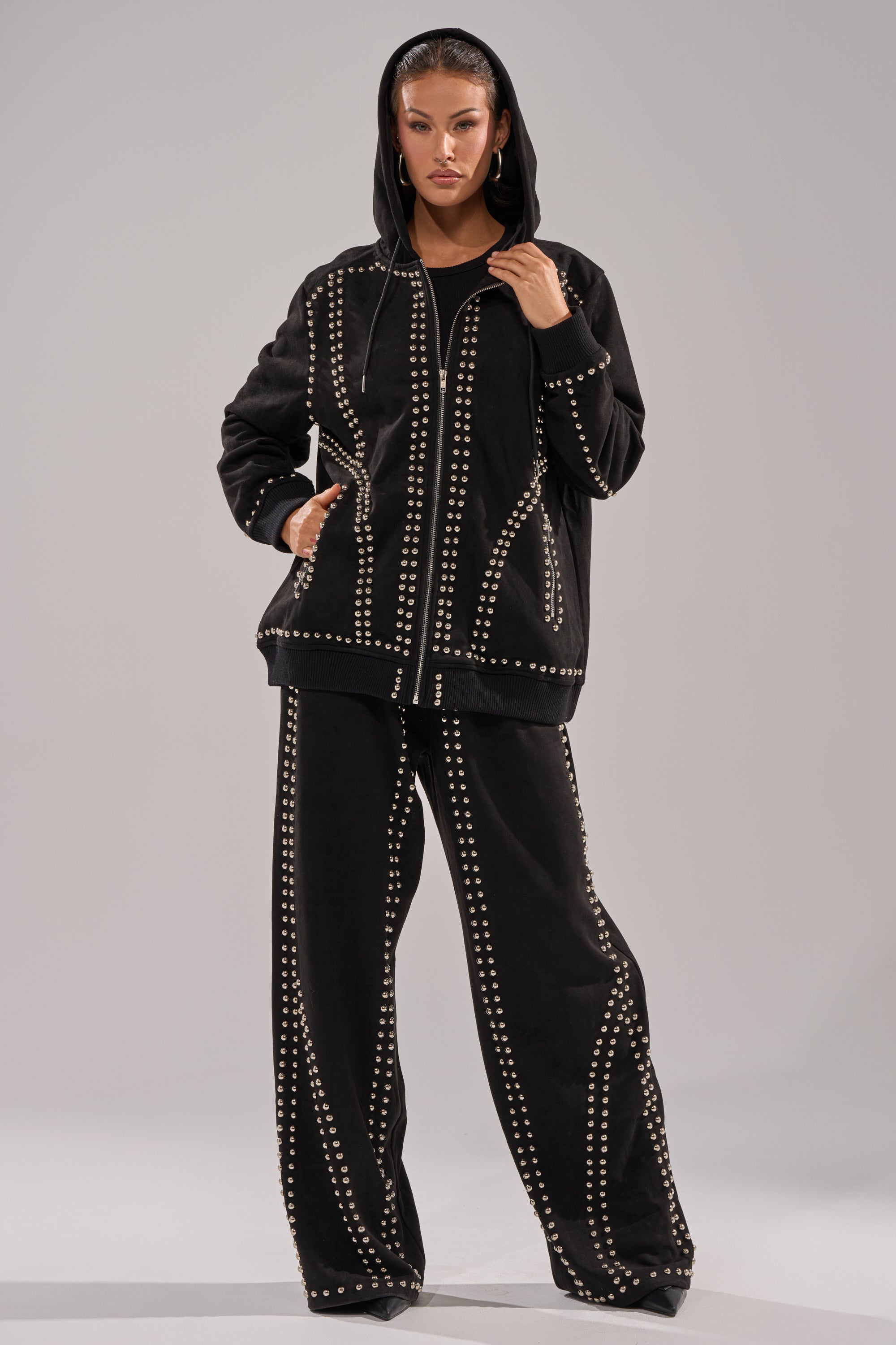 A person wears the loose-fitting ASHLEY STUDDED ZIP UP SWEATSHIRT with matching wide-leg pants, both featuring geometric silver stud details. The oversized casual outfit is shown against a plain, light background.