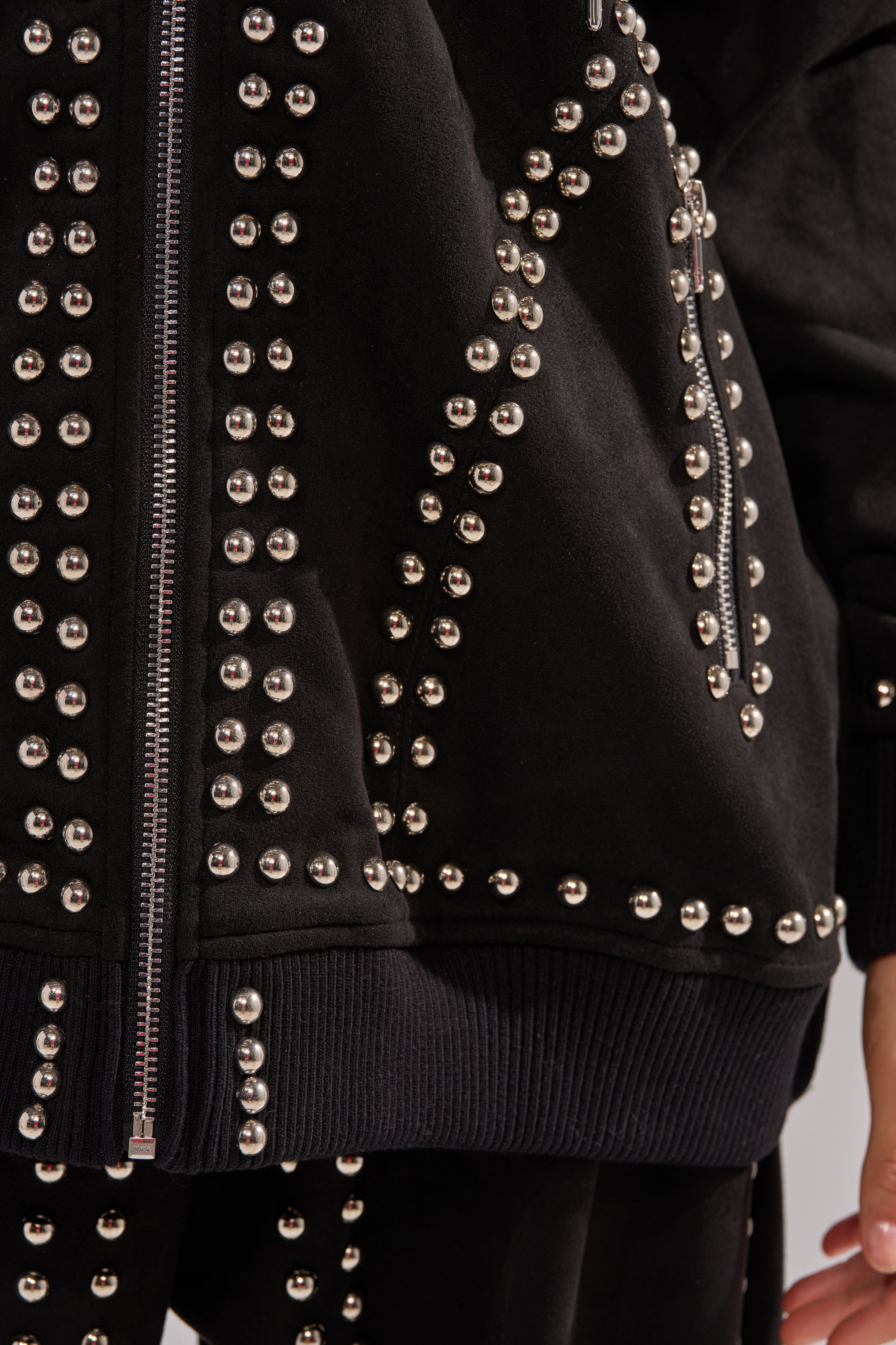 Close-up of the ASHLEY STUDDED ZIP UP SWEATSHIRT, showcasing metallic studs and zipper accents on black faux suede, with sleeve and pocket details visible along with a person’s hand.