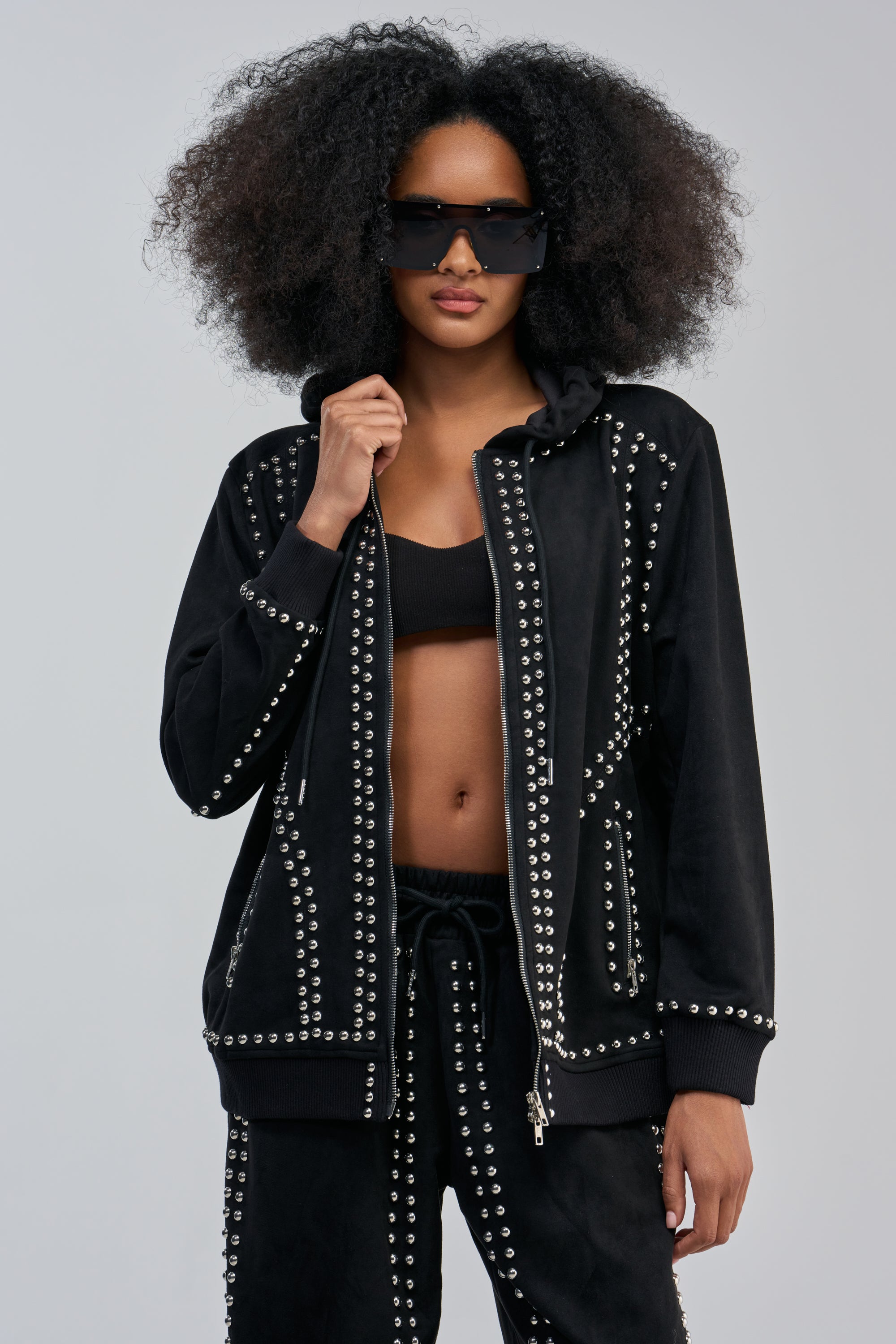 Wearing the ASHLEY STUDDED ZIP UP SWEATSHIRT, a person with a large afro and dark sunglasses poses against a light gray background. The studded black sweatshirt is paired with matching silver-studded pants, layered over a black sports bra.