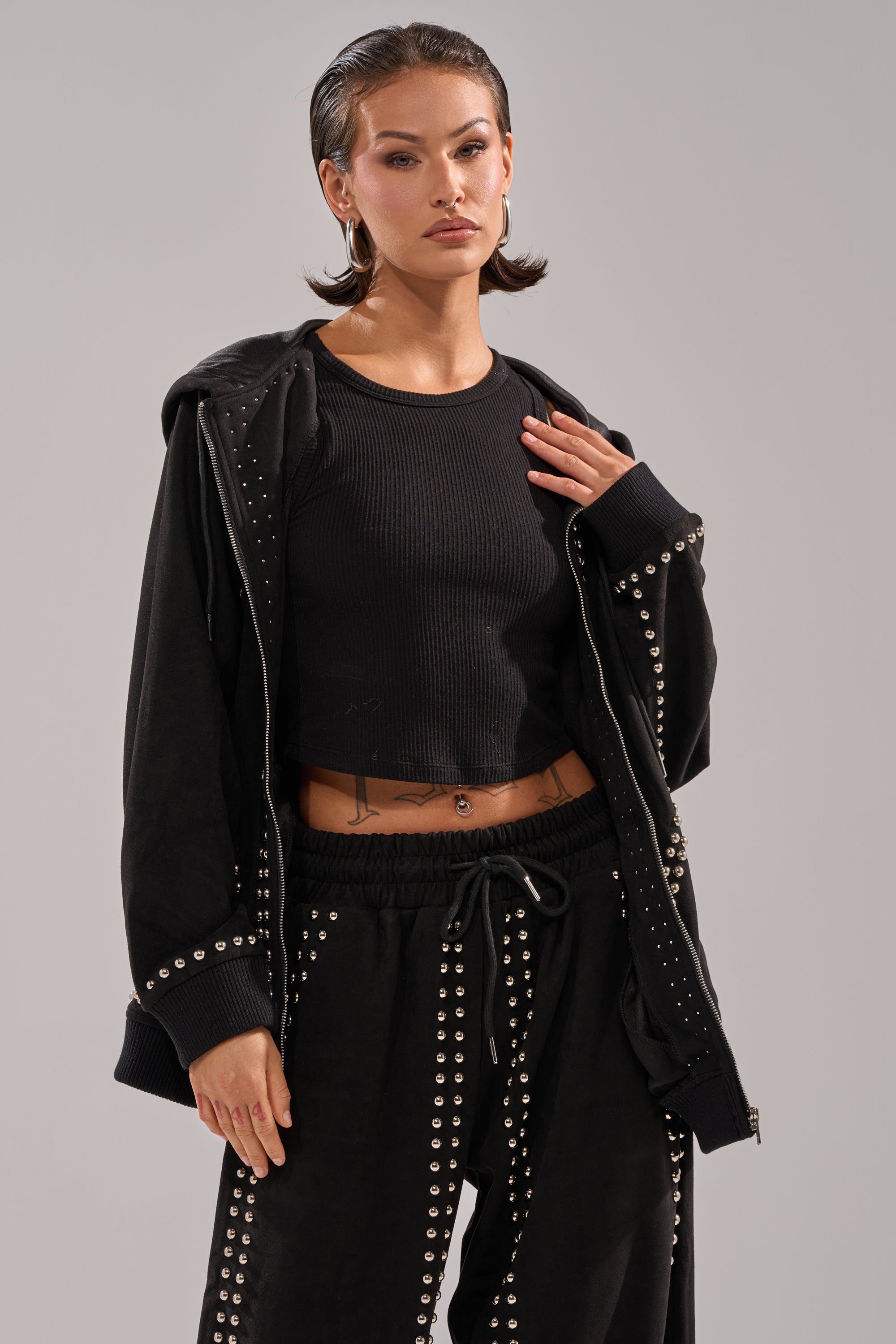 A woman with slicked-back hair wears the ASHLEY STUDDED ZIP UP SWEATSHIRT and matching studded pants over a black ribbed crop top, accessorized with hoop earrings, a belly piercing, and a tattoo above her waistband against a plain background.