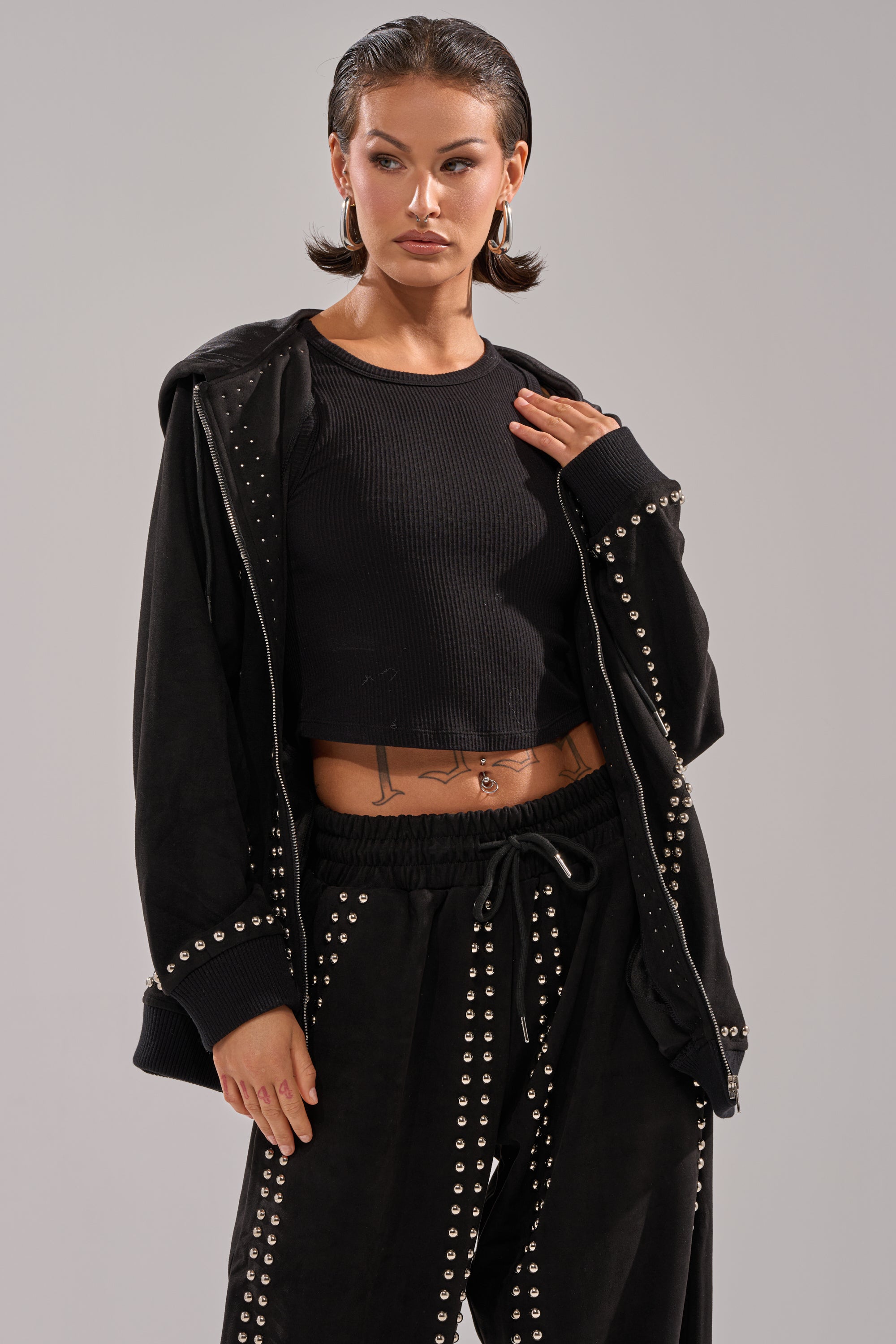 A person with short brown hair pairs a black ribbed crop top and loose studded pants with the ASHLEY STUDDED ZIP UP SWEATSHIRT draped over their shoulders, revealing a tattoo on their midriff.