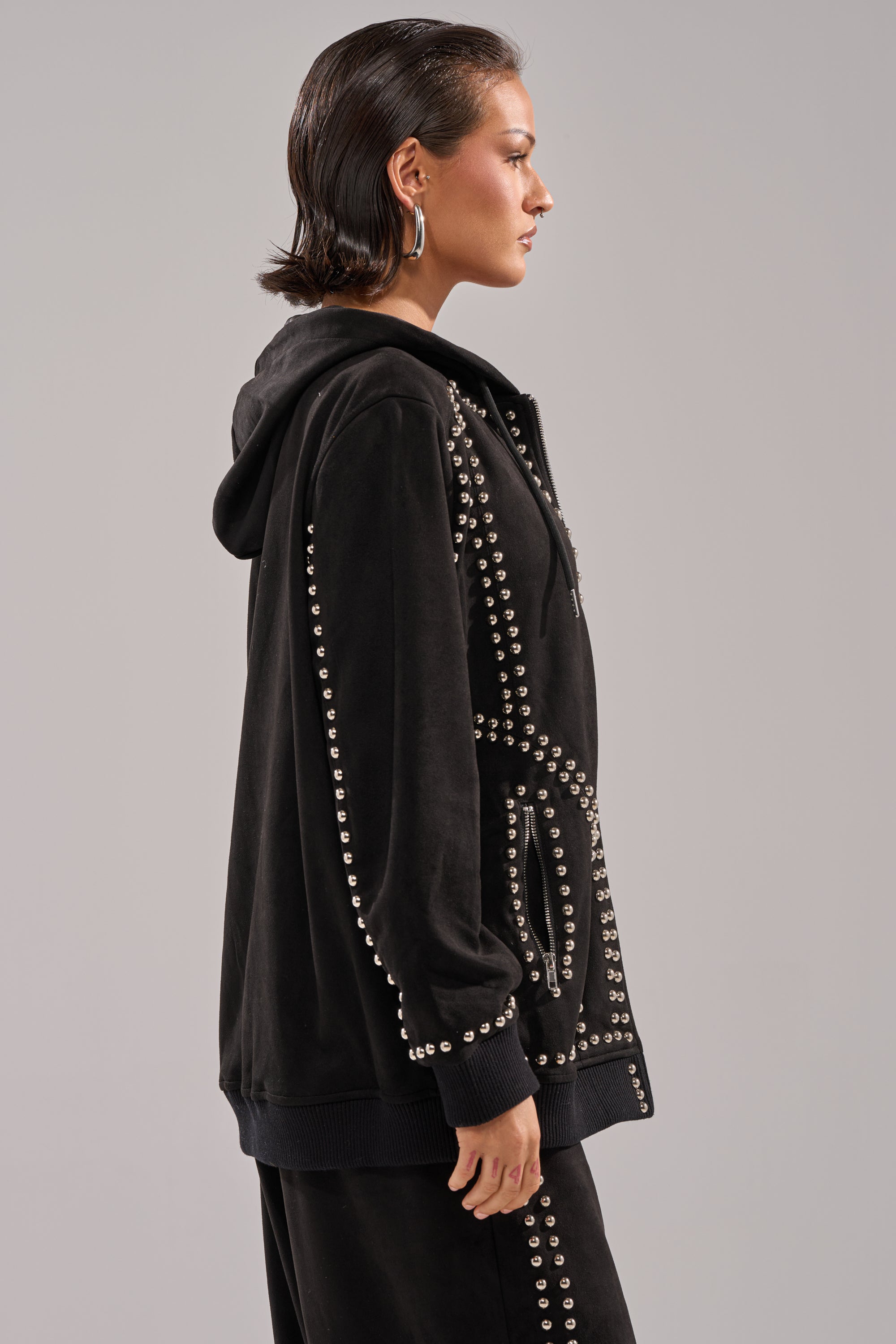 A woman with slicked-back dark hair models the ASHLEY STUDDED ZIP UP SWEATSHIRT and matching black pants, accessorized with large hoop earrings, standing in profile against a neutral gray background.