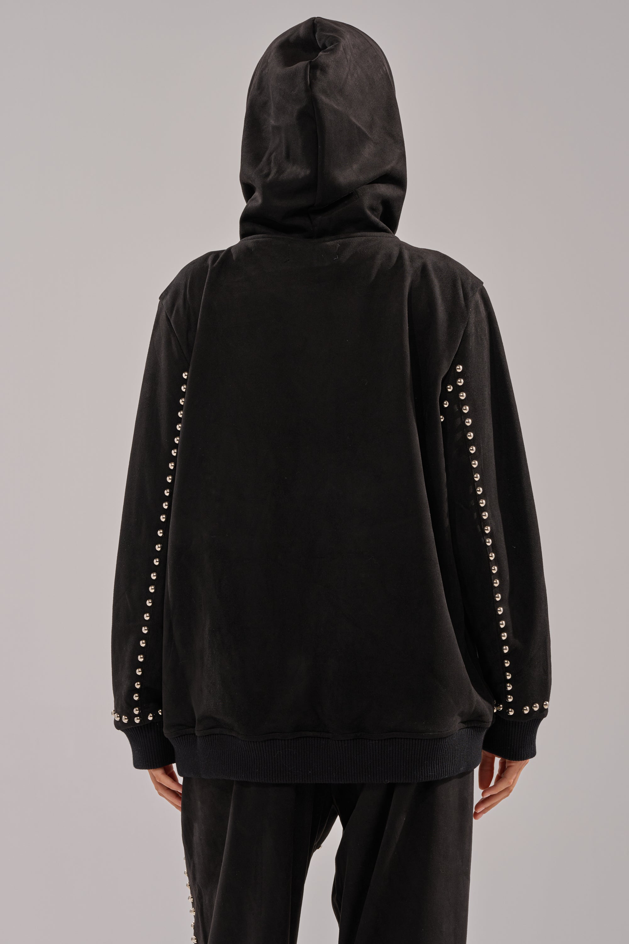 A person is shown from the back wearing the ASHLEY STUDDED ZIP UP SWEATSHIRT, a black sweatshirt with silver studs along the sleeves, paired with matching black pants.