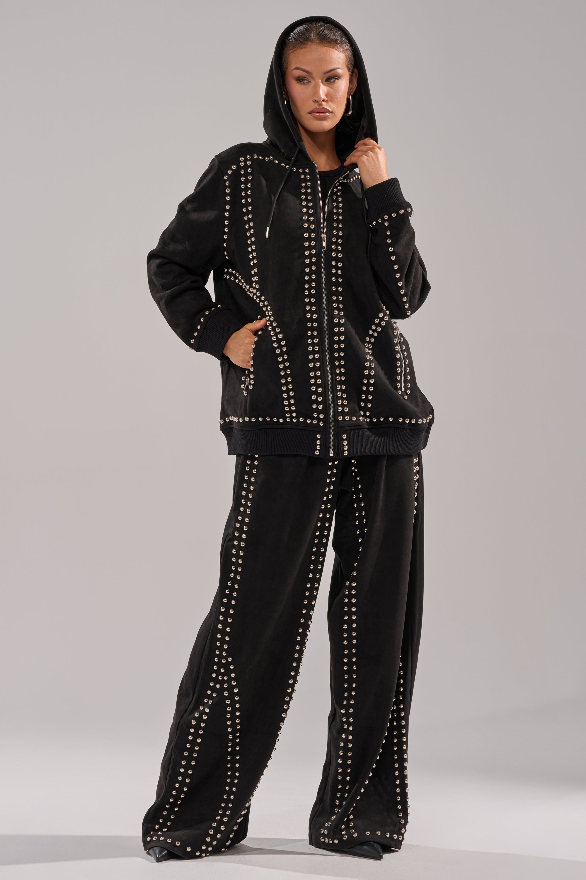 A woman confidently poses in the ASHLEY STUDDED ZIP UP SWEATSHIRT with matching wide-leg pants, featuring silver studded embellishments—one hand on her hood, the other in her pocket.