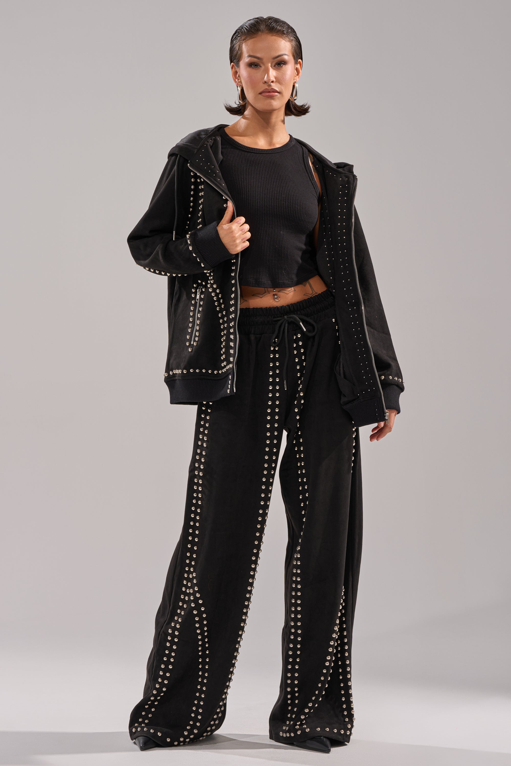 A woman stands confidently in a black ribbed crop top, wide-leg pants, and the ASHLEY STUDDED ZIP UP SWEATSHIRT—an oversized zip up featuring rows of metal studs—set against a plain gray background.
