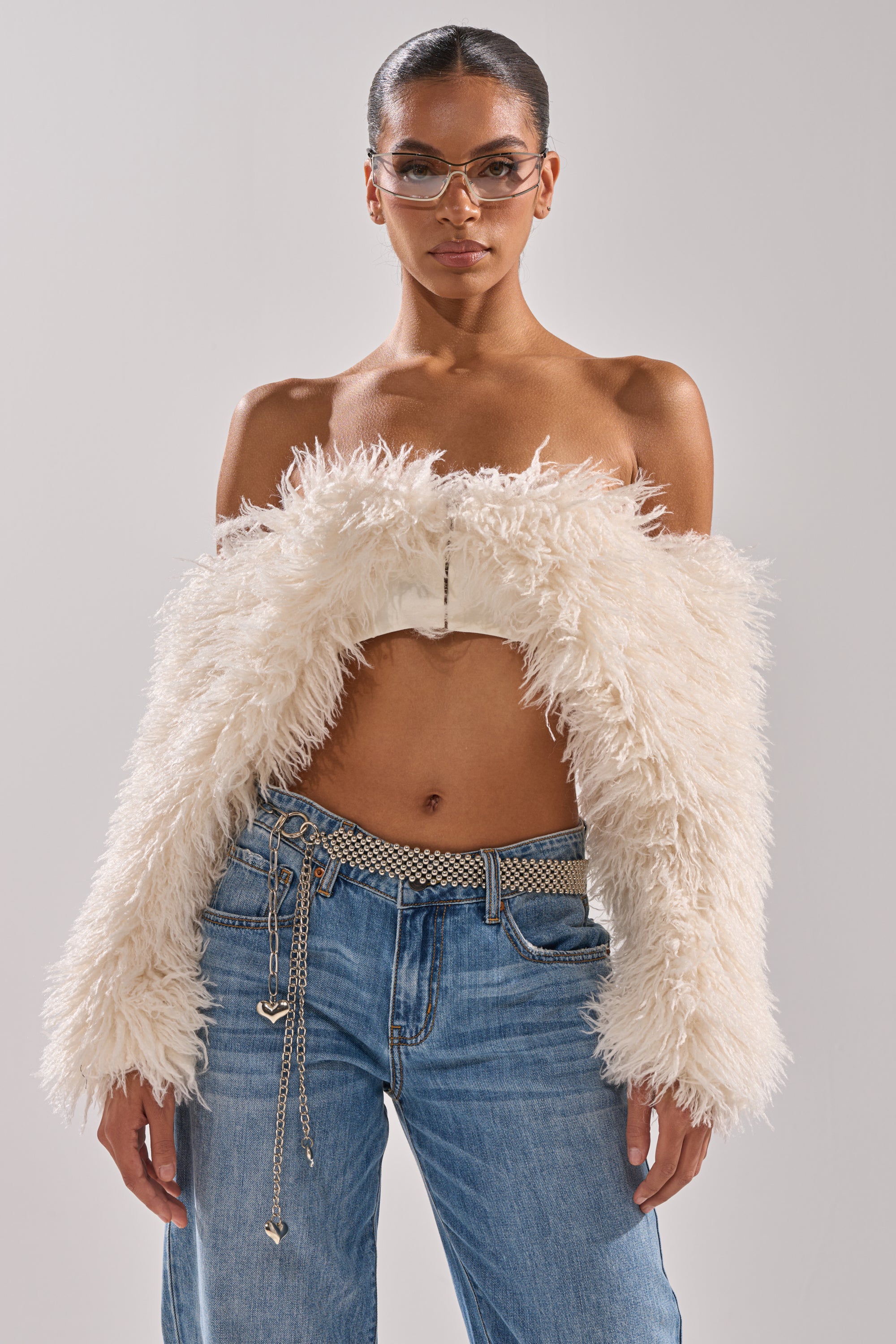 A woman wearing the SNOW FUR TOP off the shoulder with clear glasses and a metallic chain belt on blue jeans stands confidently against a neutral background. 0