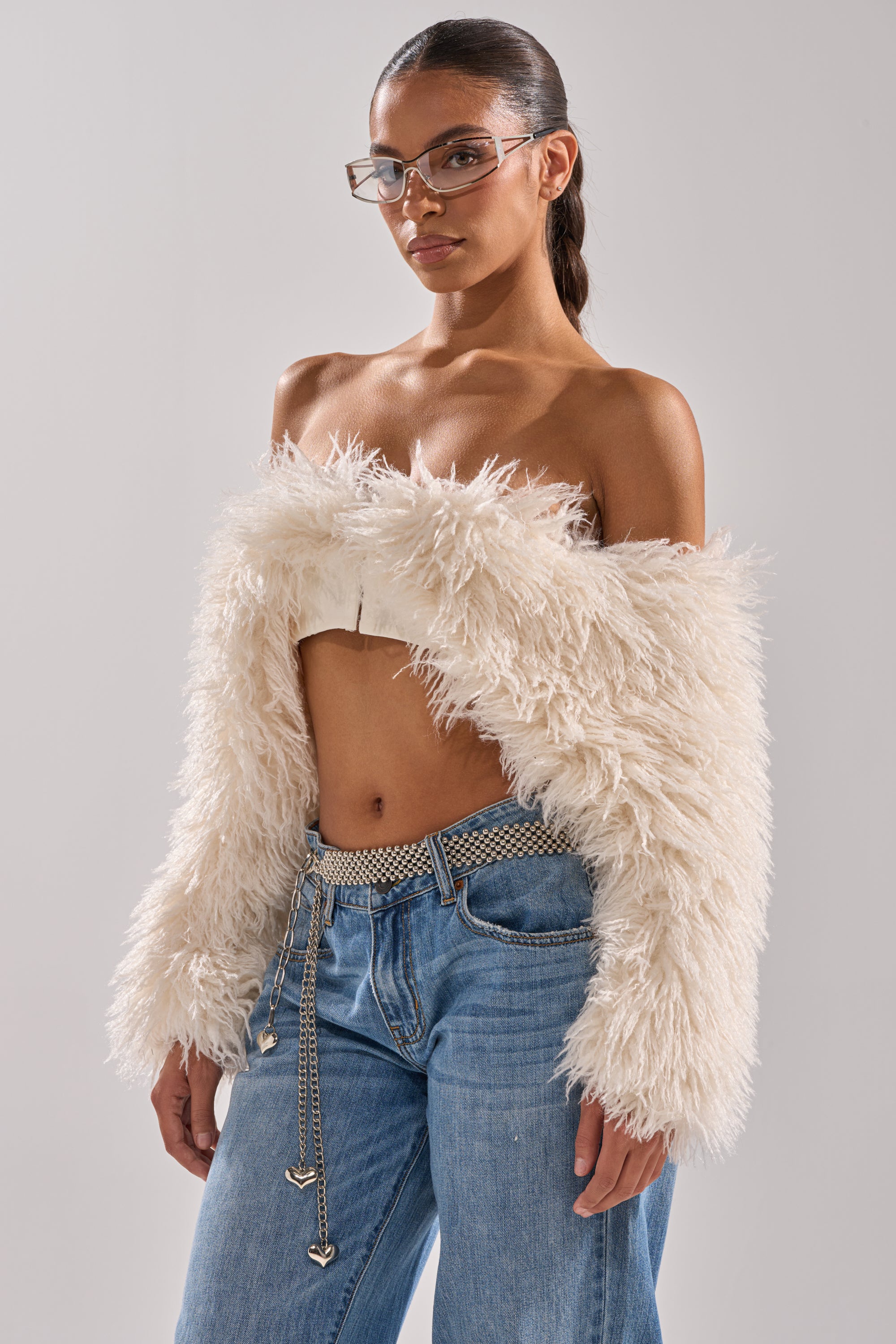 A person wearing the SNOW FUR TOP, blue jeans with a silver chain belt, and glasses stands against a plain light grey background. 1