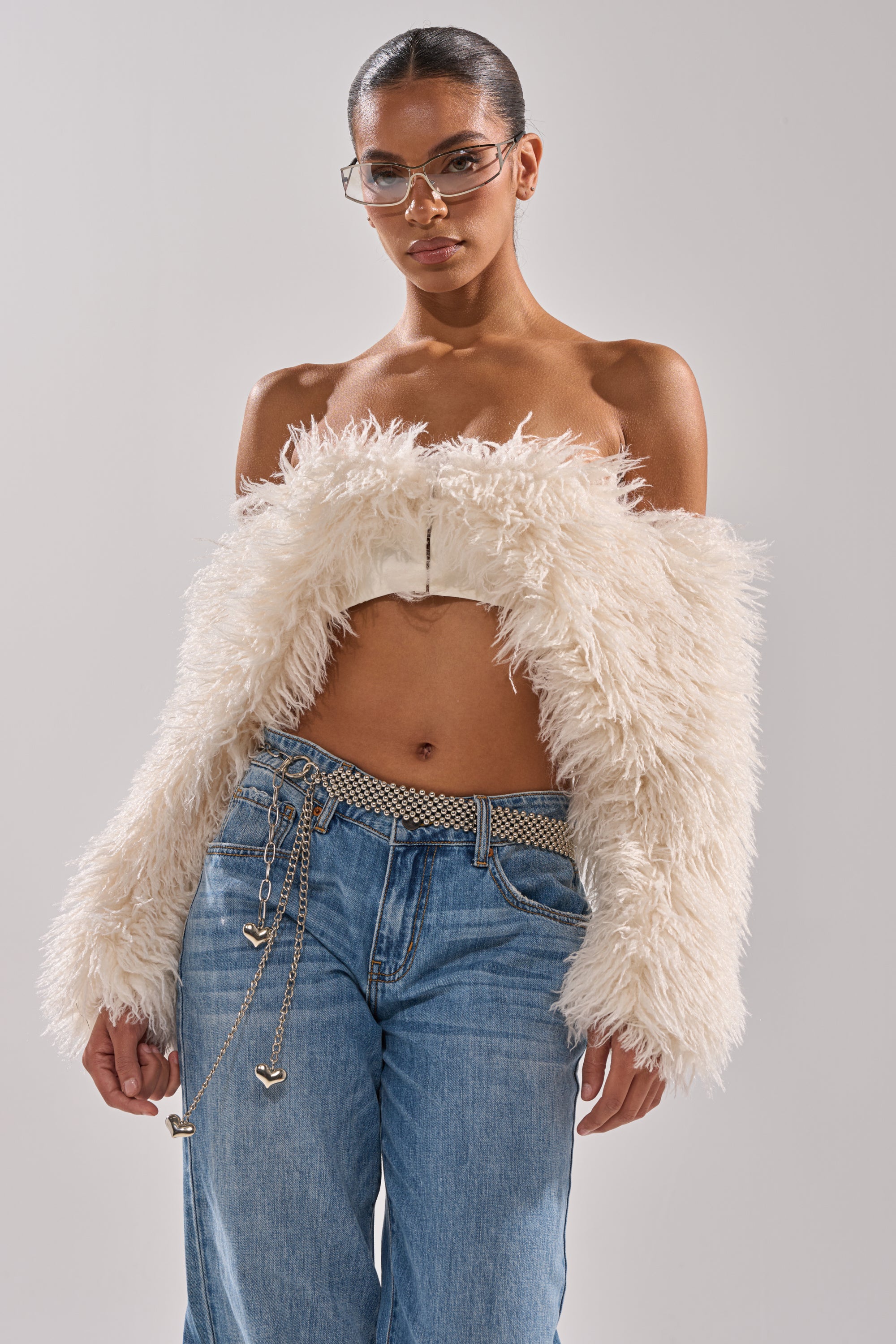 Wearing the SNOW FUR TOP off-shoulder cream jacket, light blue jeans with a silver chain belt, and sunglasses, a woman stands confidently against a plain background with a sleek hairstyle. 2