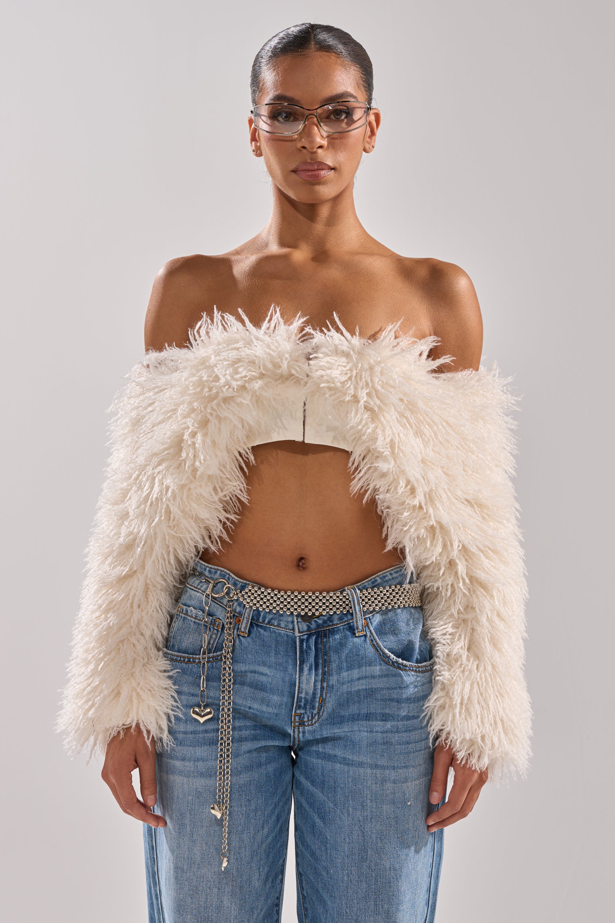 A woman in glasses wears the SNOW FUR TOP, a white cropped off-the-shoulder piece with a fur texture, paired with blue jeans and a chain belt with heart charms, standing against a plain background. 3