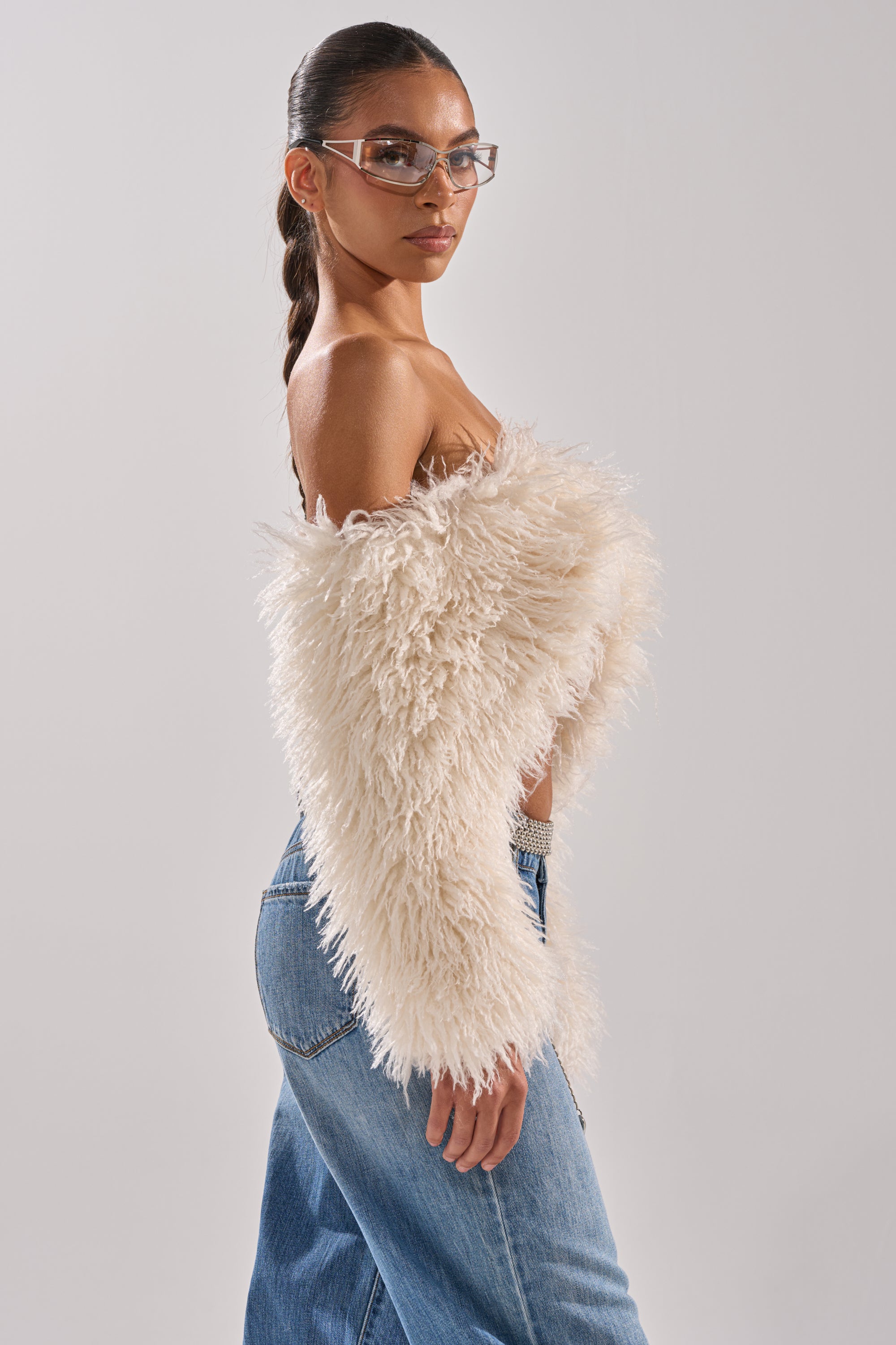 A woman with long, dark hair in a ponytail wears clear glasses, a SNOW FUR TOP, and blue jeans, standing in profile against a plain light background. 4