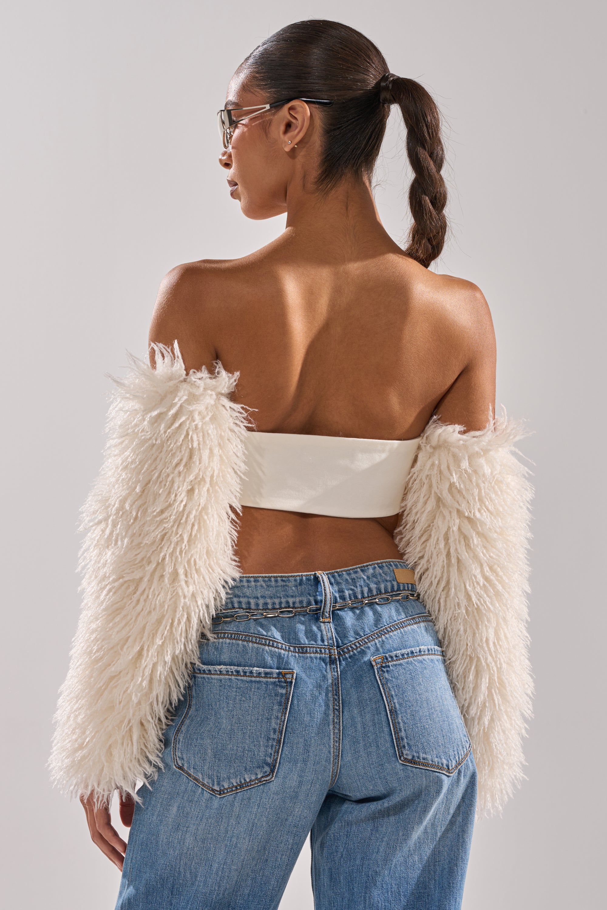 A woman in sunglasses and a sleek braided ponytail models the SNOW FUR TOP—an off-shoulder jacket—paired with high-waisted blue jeans, posing with her back to the camera against a plain background. 5