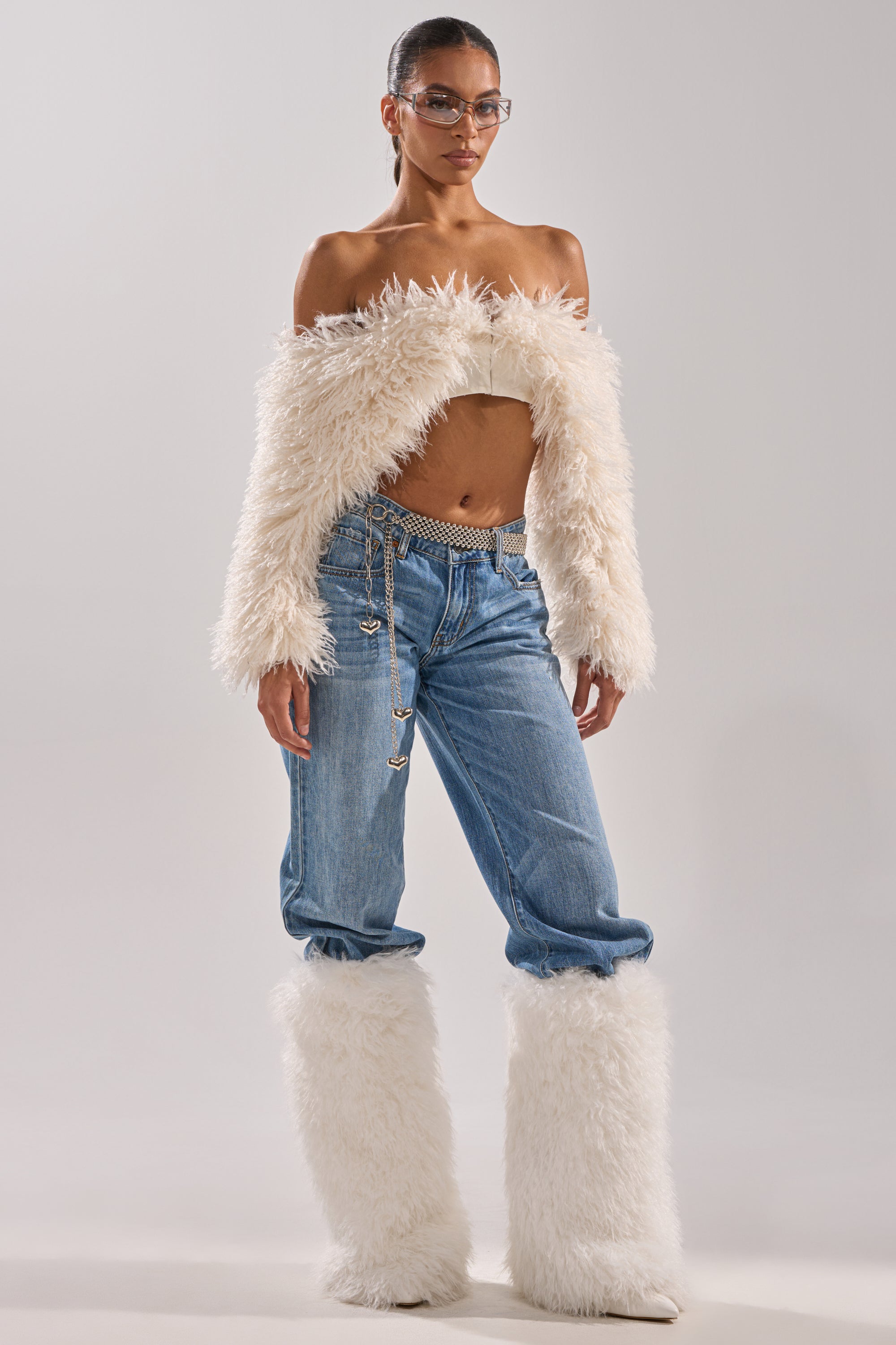 A woman in a SNOW FUR TOP off-the-shoulder jacket, wide-leg blue jeans with a chain belt, matching fluffy white leg warmers, and clear glasses stands against a plain light background. 8