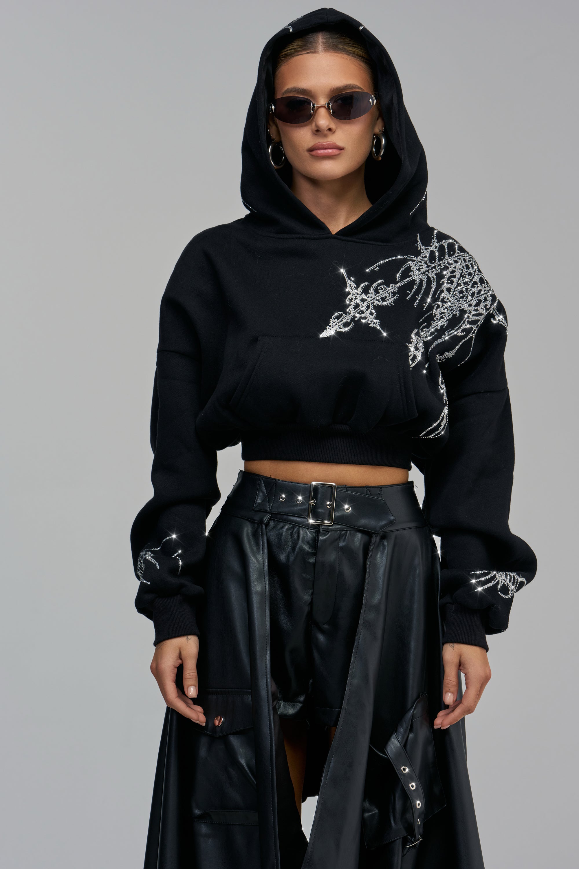 A person wears a black LEXI CROPPED RHINESTONE HOODIE with silver rhinestone embellishments on the shoulders and sleeves, styled with black pants for a grunge streetwear look.