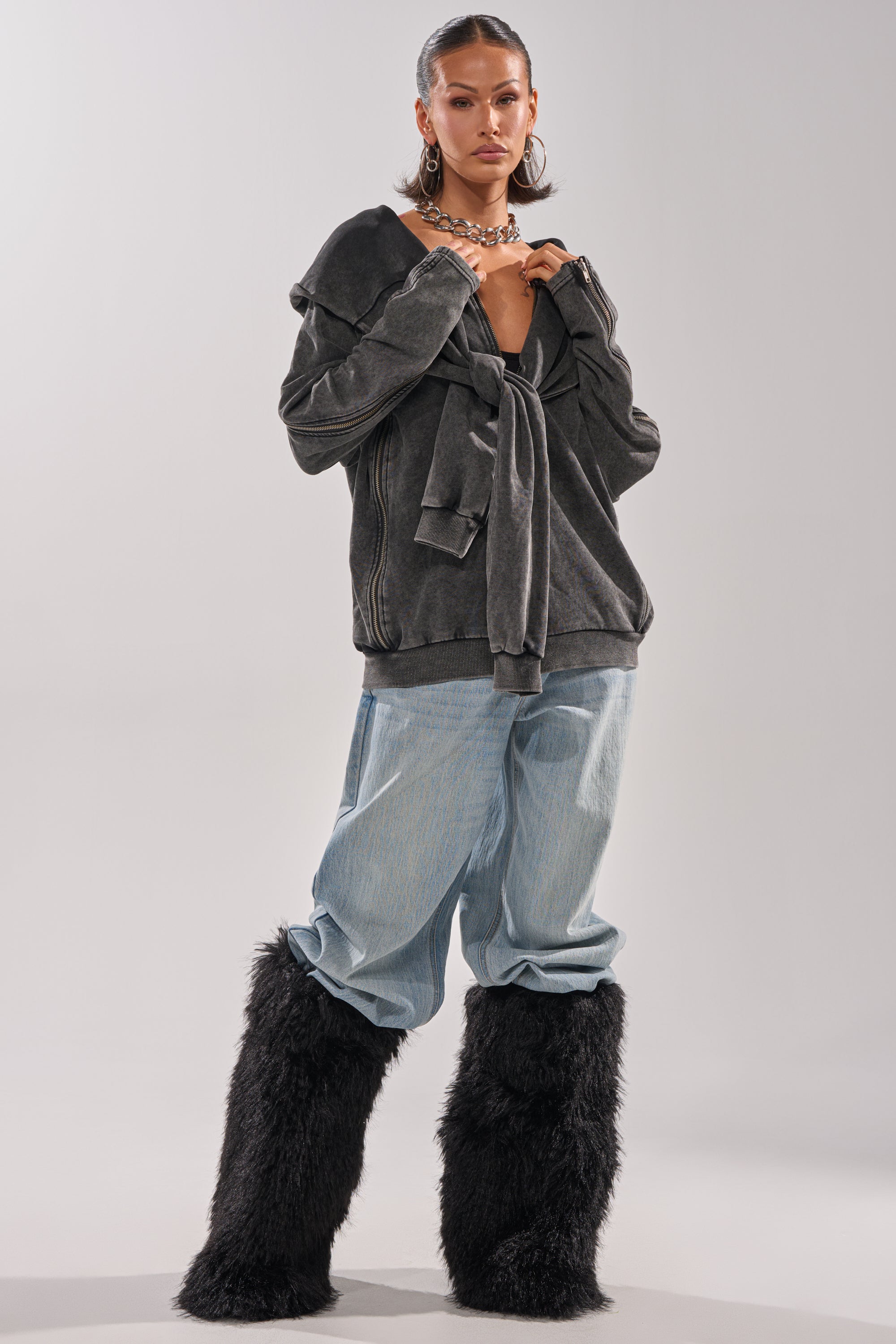 A woman models the COOL GAL TIE SWEATSHIRT in dark gray, pairing it with light blue baggy jeans and black fluffy knee-high boots. She completes her elevated loungewear look with hoop earrings, a chunky necklace, and sleek hair.