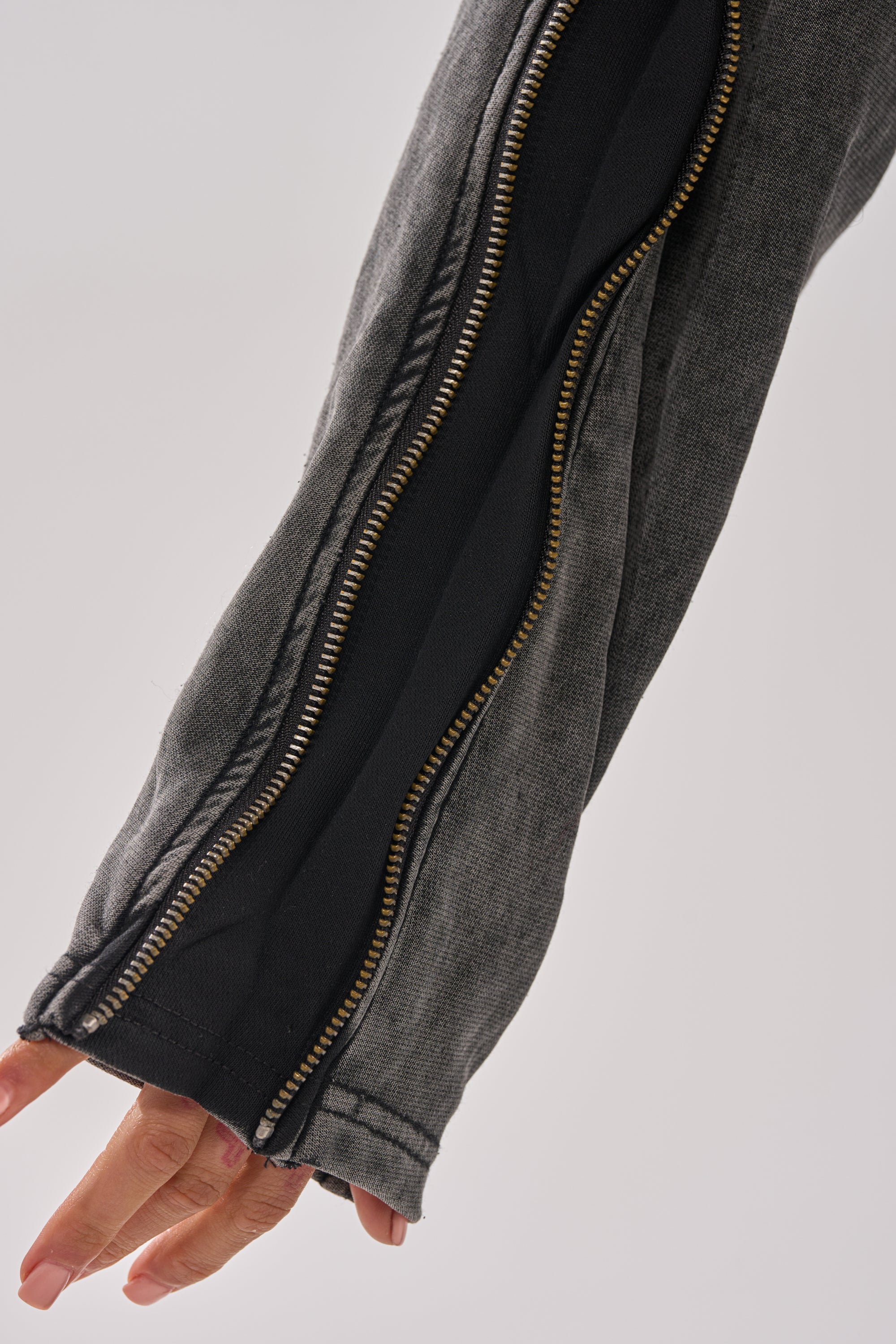 A close-up of a person's arm in the COOL GAL TIE SWEATSHIRT, featuring a gray denim sleeve with exposed brass zippers over a charcoal hoodie. The person's hand peeks out from the end of the sleeve.