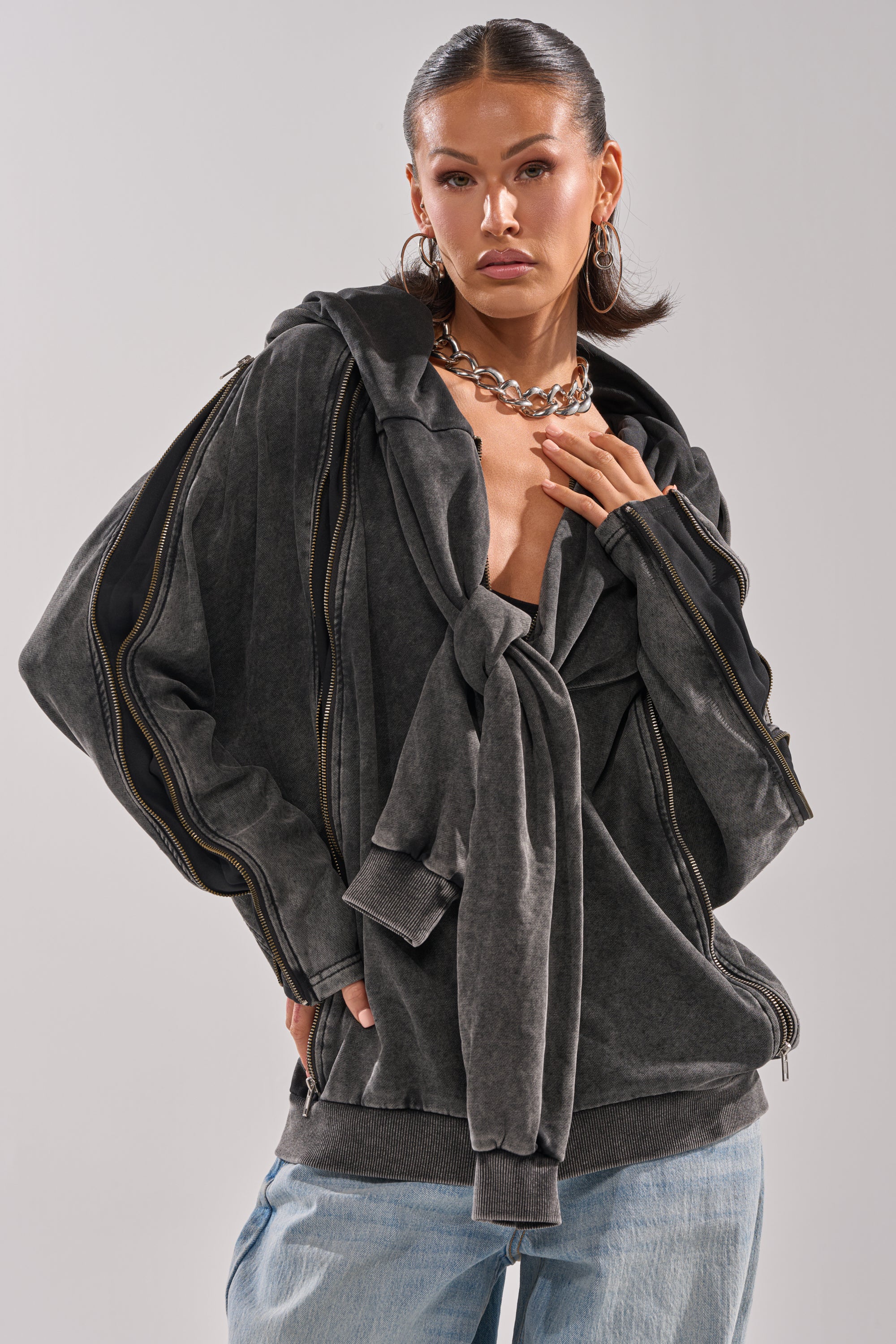 A woman with slicked-back hair wears the COOL GAL TIE SWEATSHIRT—charcoal with oversized sleeves and a large front tie—paired with light blue jeans, hoop earrings, and a chunky chain necklace, standing against a neutral background.