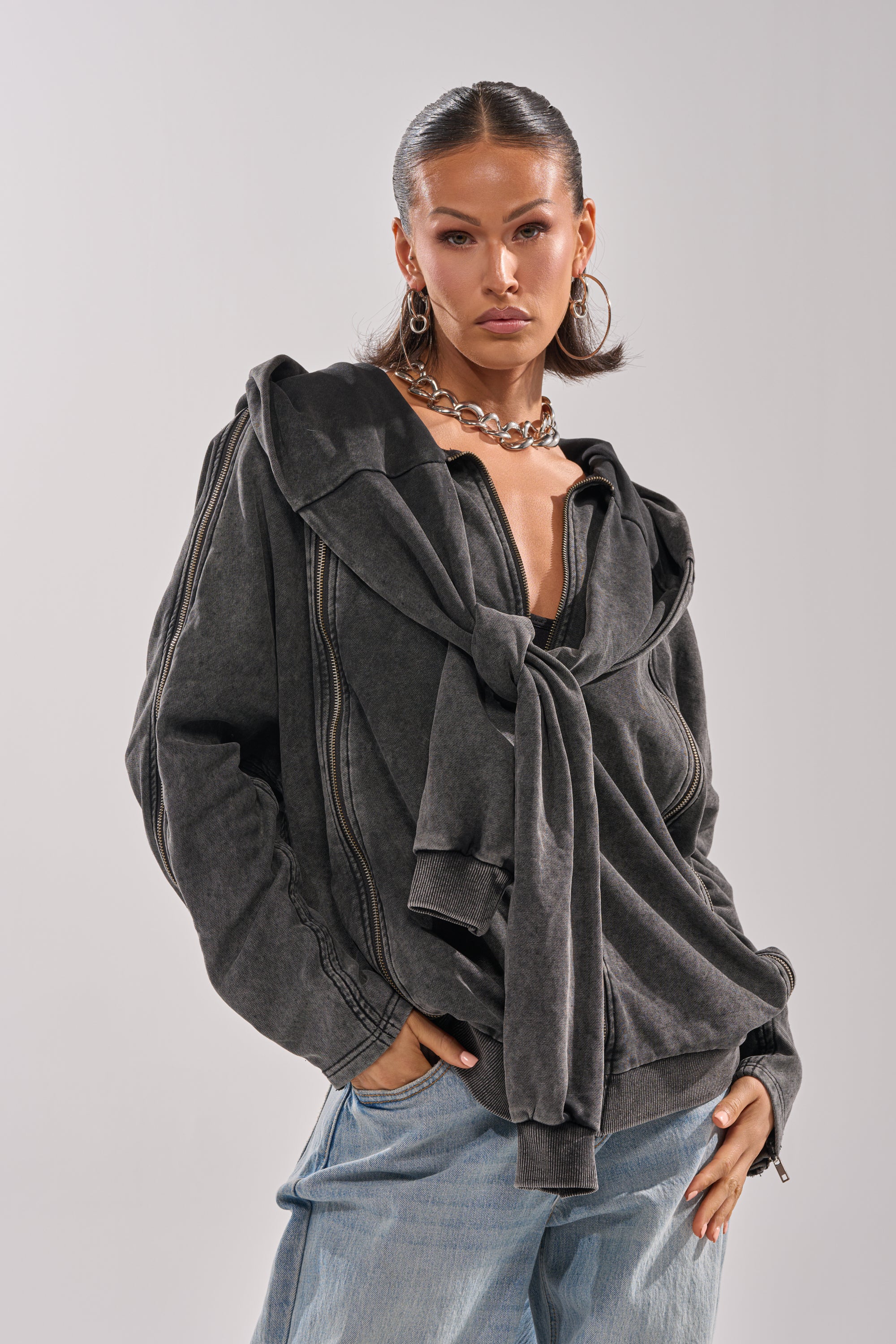 A woman with slicked-back hair layers the COOL GAL TIE SWEATSHIRT over a dark oversized cotton knit sweat, pairing it with light blue jeans, large hoop earrings, and a chunky chain necklace for an elevated loungewear look.