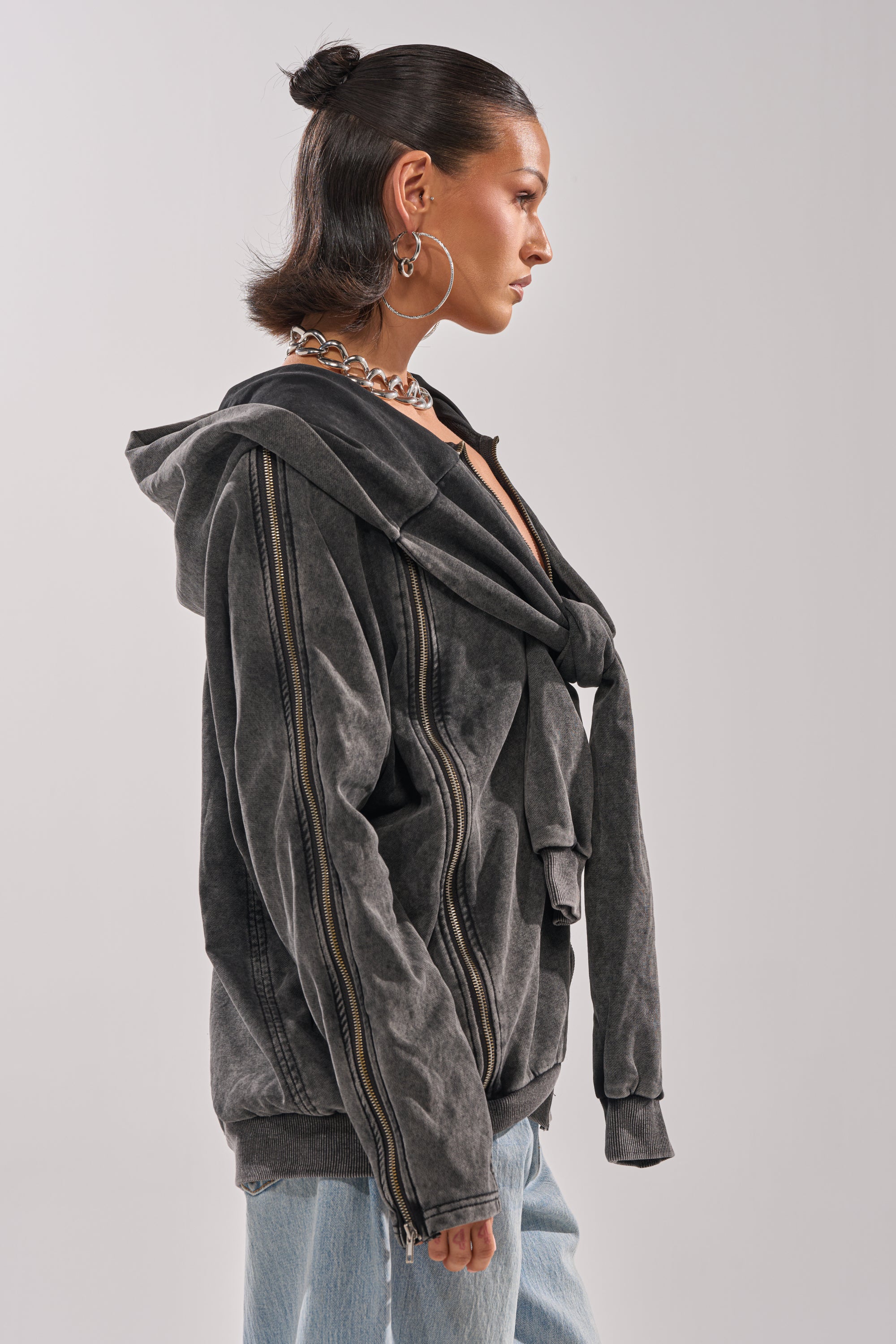 A woman with dark slicked-back hair, hoop earrings, and a chunky chain necklace models the COOL GAL TIE SWEATSHIRT—a loose charcoal hoodie with an extra attached sleeve detail—over light blue jeans for a relaxed loungewear look.