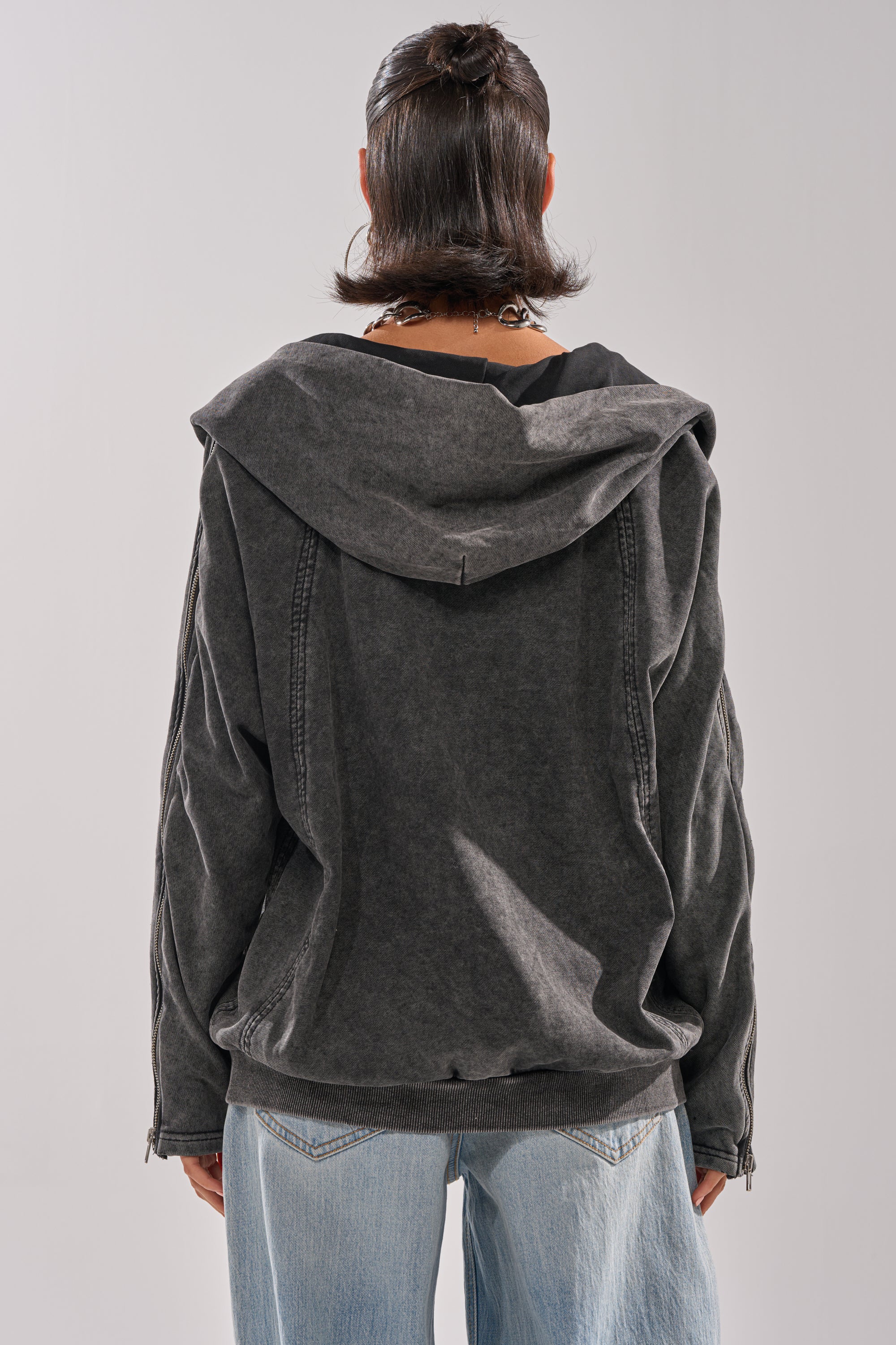 A person with shoulder-length dark hair is seen from behind, wearing the COOL GAL TIE SWEATSHIRT in black with a hood and light blue jeans, against a plain light background.