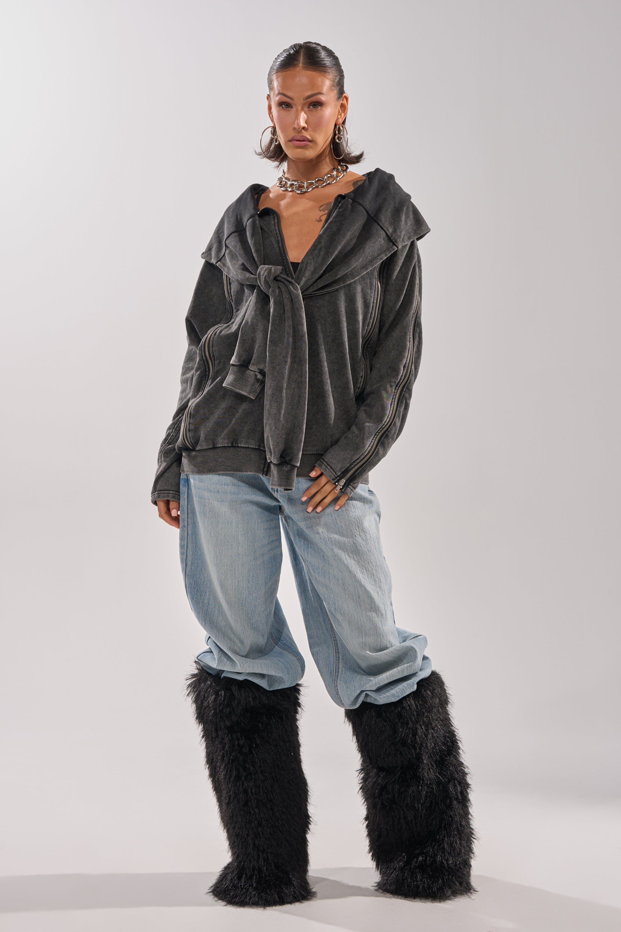 A woman wears the COOL GAL TIE SWEATSHIRT in charcoal, paired with light blue baggy jeans, chunky chain necklaces, and tall black furry boots, standing against a plain light background.