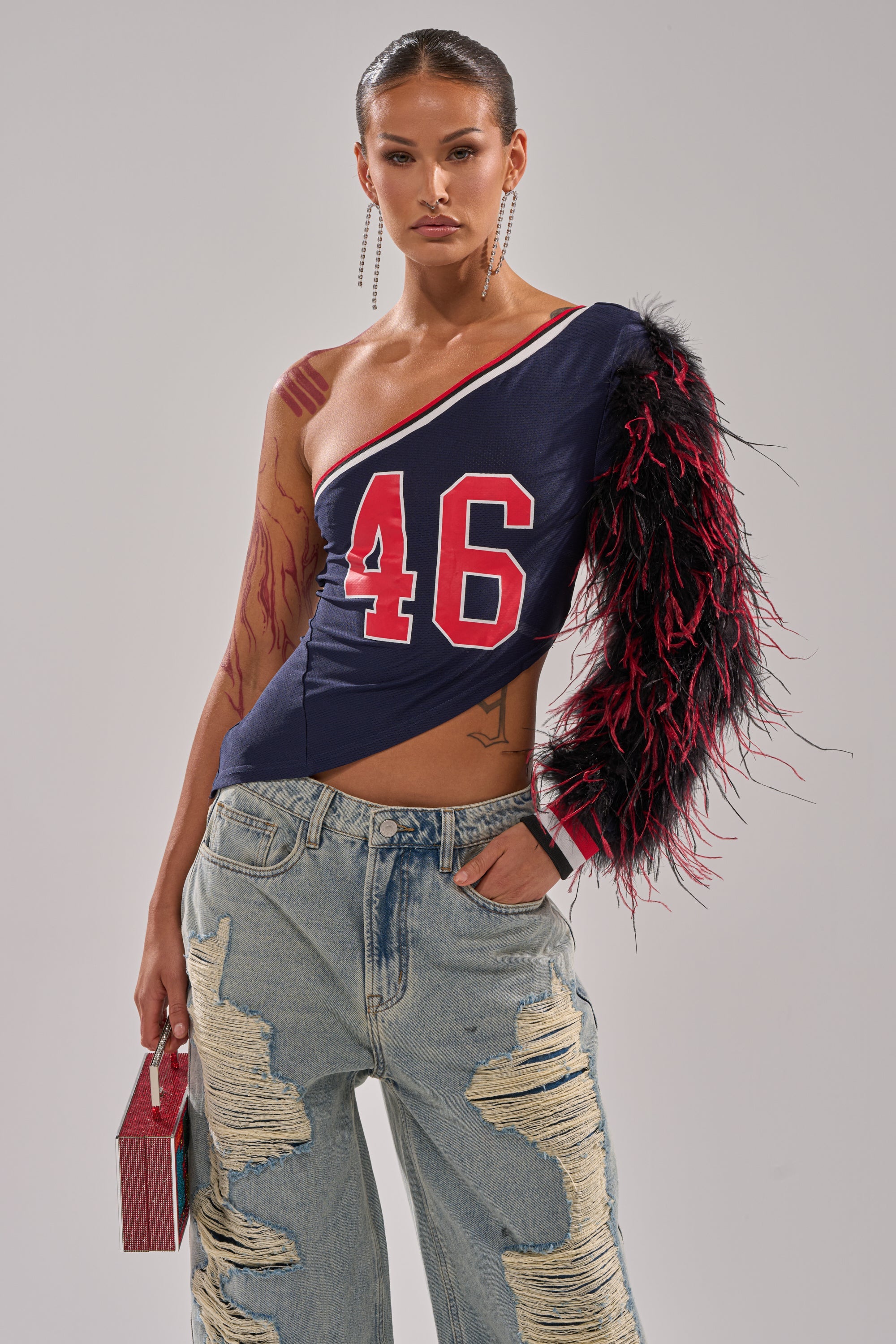 A woman models the navy SUPERBOWL PARTY ONE SLEEVE TEE with feathered sleeve and number 46, one hand in her ripped jeans pocket. She carries a small red purse, wears long earrings, and has slicked-back hair. 0