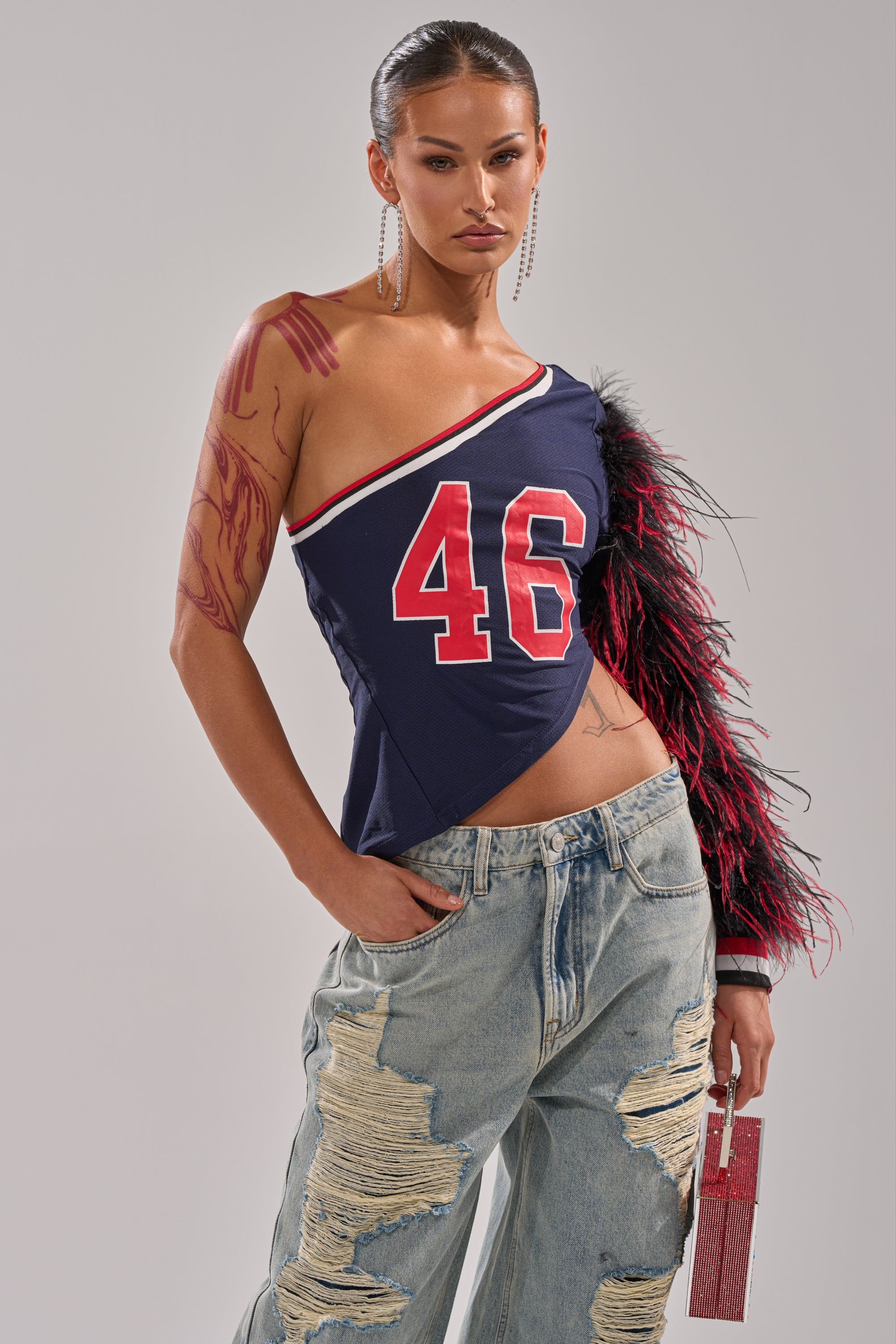 A woman wears the SUPERBOWL PARTY ONE SLEEVE TEE—a navy one-shoulder top with a red "46" graphic and black and pink feathered sleeve—paired with ripped baggy jeans, long earrings, and a small pink handbag. 1