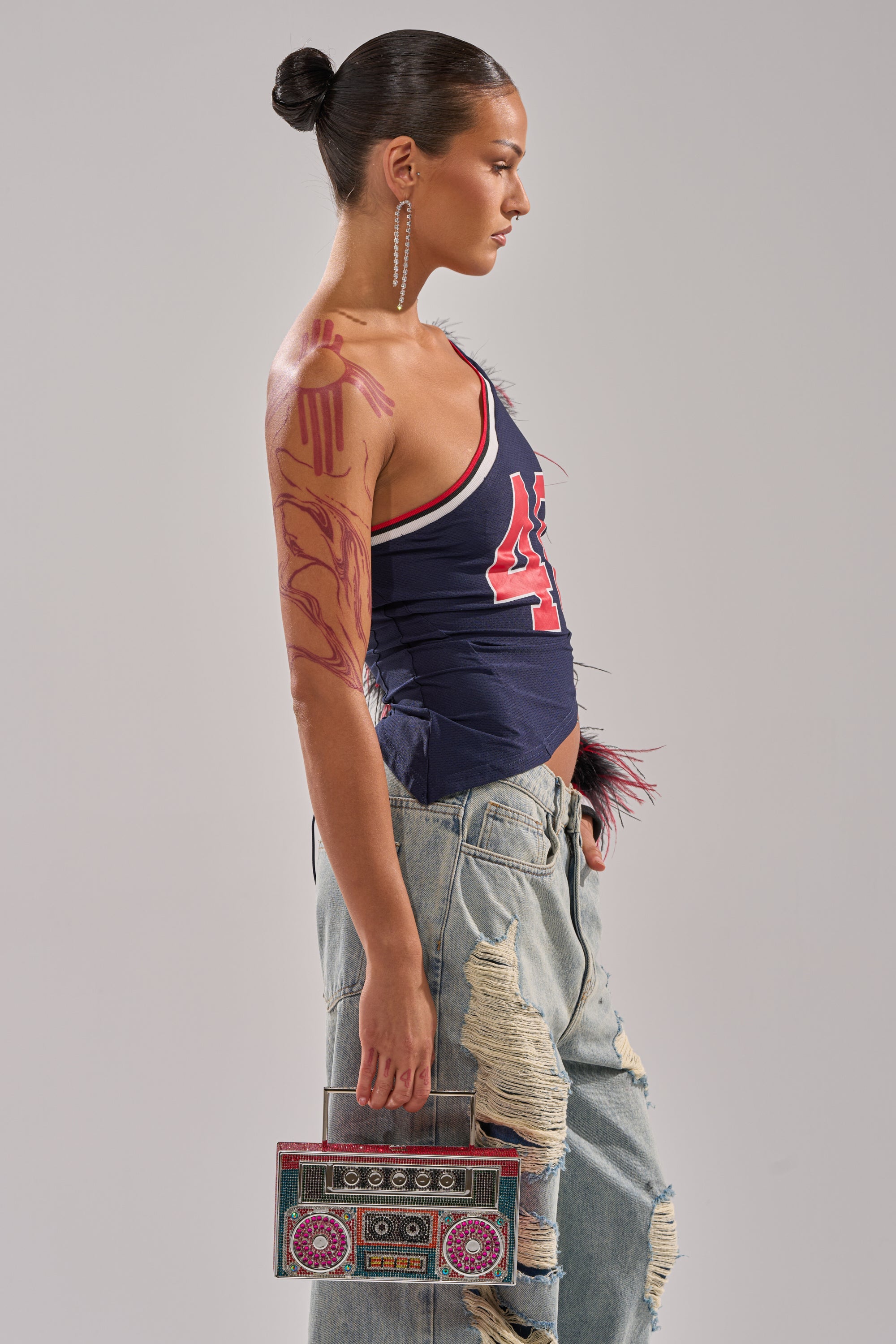 A woman with slicked-back hair stands in profile, wearing the SUPERBOWL PARTY ONE SLEEVE TEE and ripped jeans. She holds a colorful boombox-shaped handbag and has a bold red design painted on her arm. 2