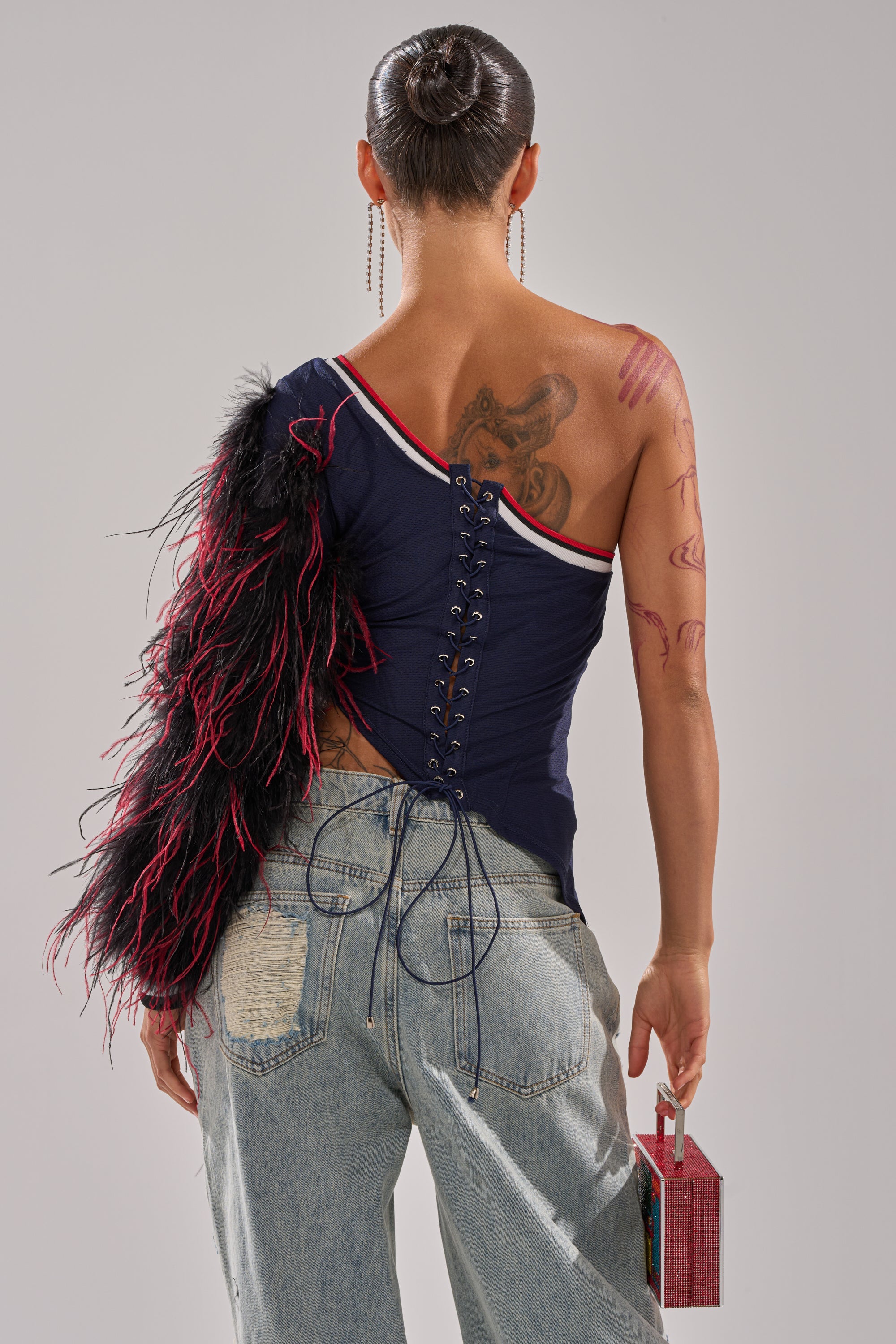 A person stands facing away, wearing the SUPERBOWL PARTY ONE SLEEVE TEE with a corset back and feathered sleeve, light blue distressed jeans, hoop earrings, holding a small pink handbag. Their hair is in a bun with visible tattoos. 3